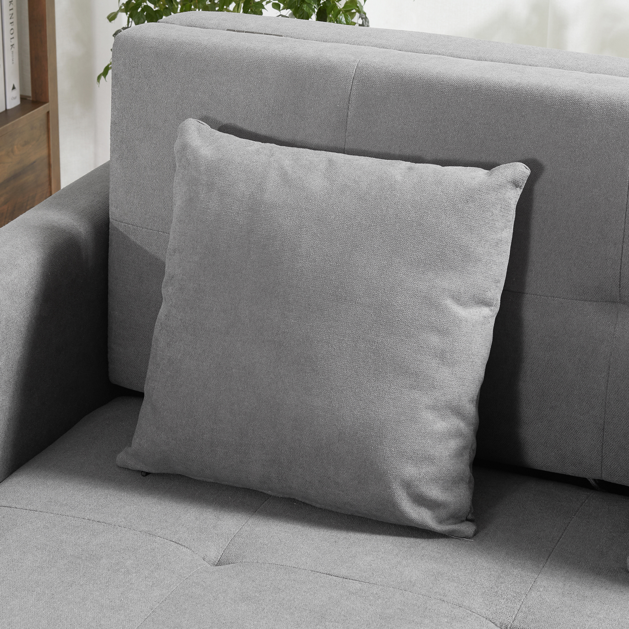 Una almohada gris cuadrada descansa sobre un sofá gris. El sofá y la almohada son de tela. Una planta está al fondo.