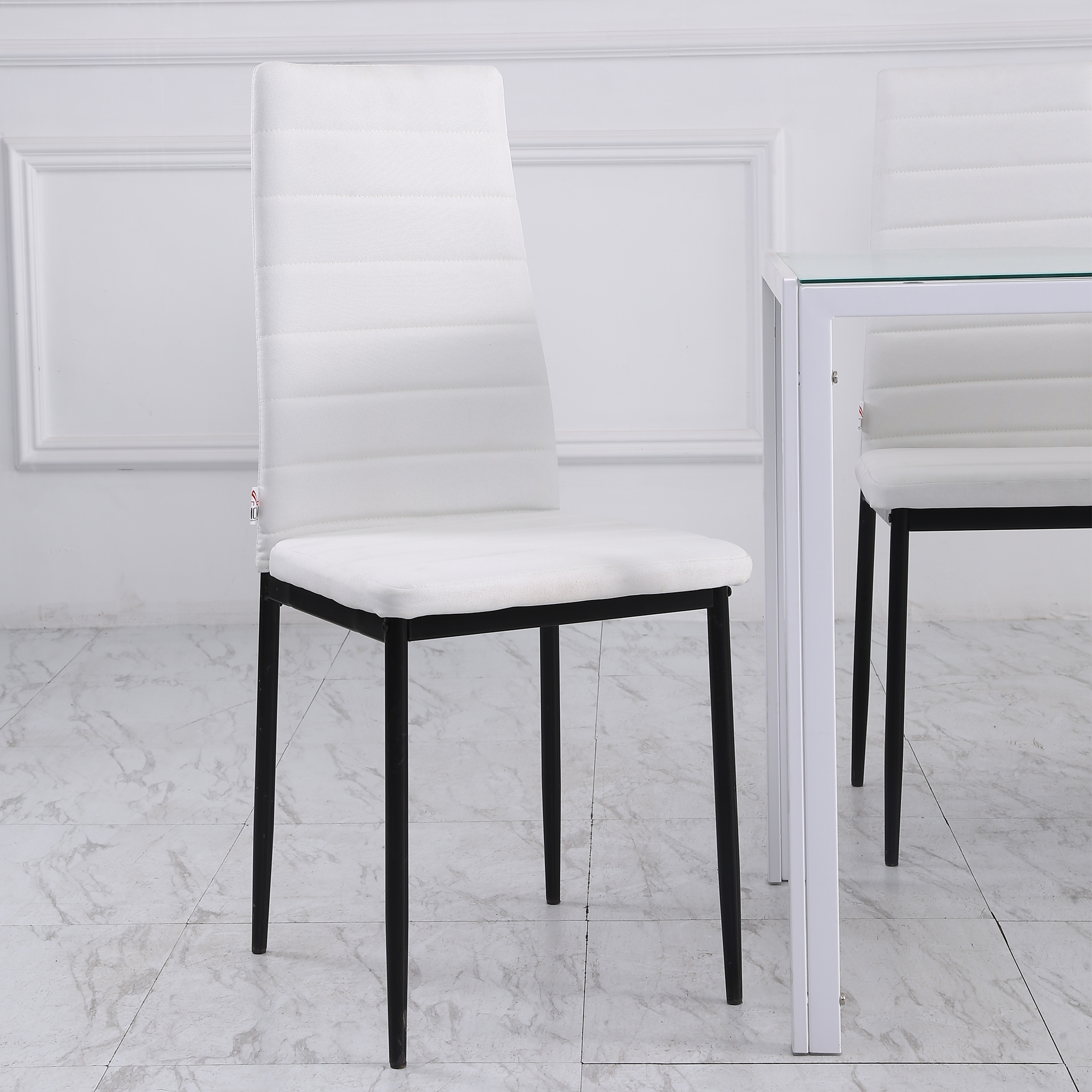 Silla blanca acolchada con patas negras. Pata de la mesa visible. Fondo: pared blanca y suelo de mármol.