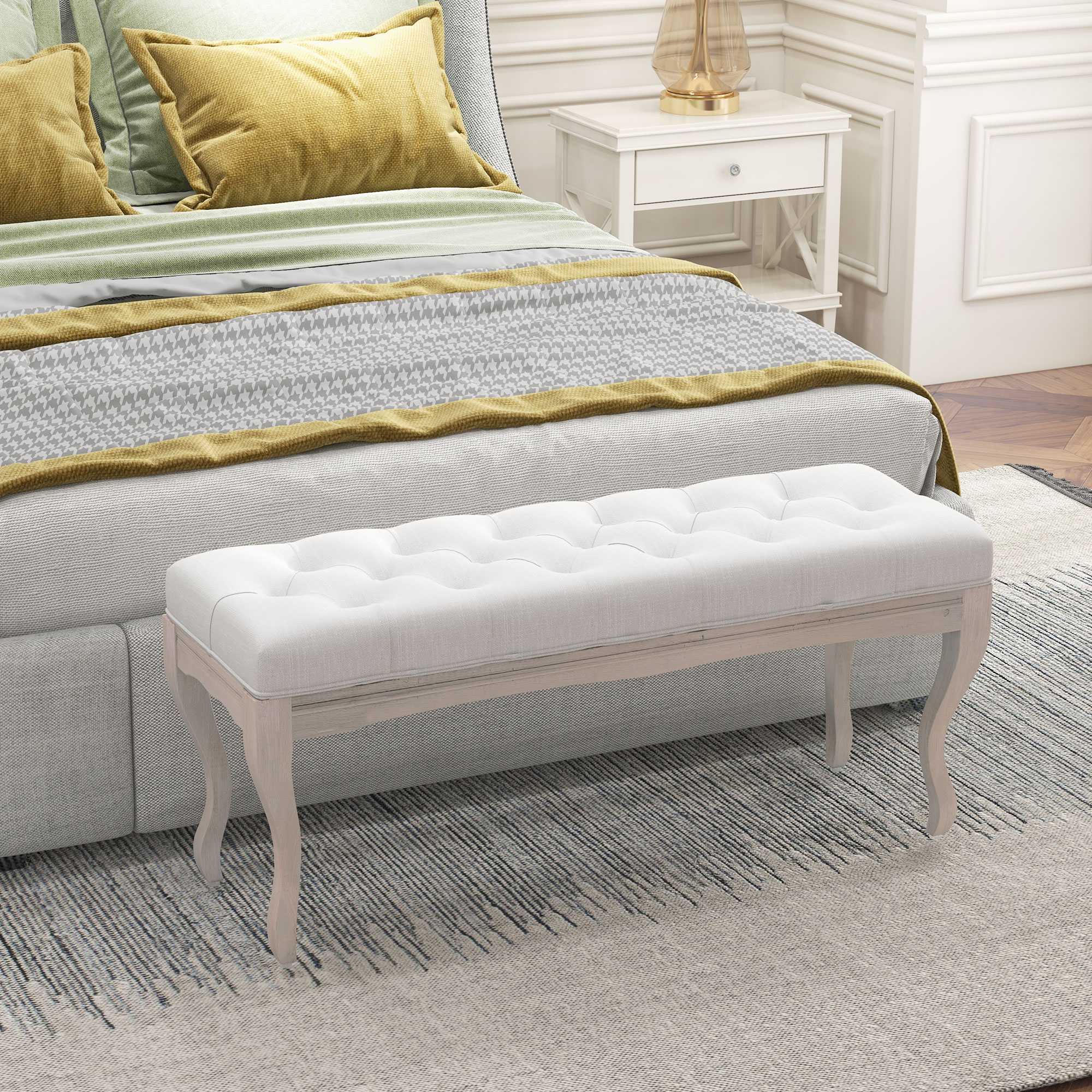 Escena de dormitorio con cama, banco, alfombra y mesita de noche. La cama tiene almohadas amarillas y una manta estampada.
