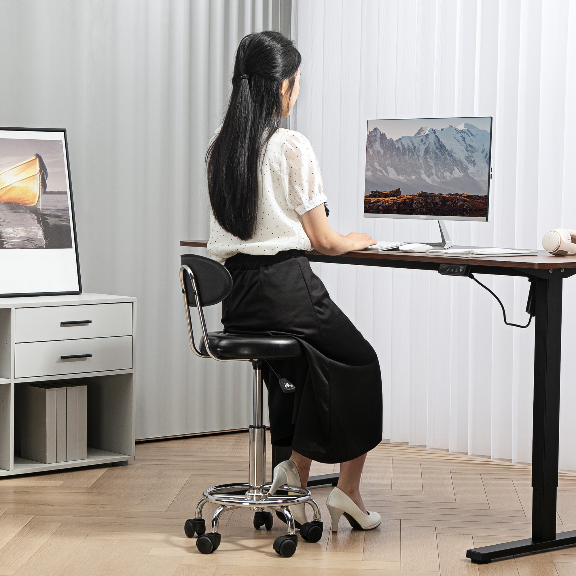 Una mujer está sentada en un escritorio, frente a una computadora. El escritorio tiene un paisaje de montaña en la pantalla. Viste pantalón negro y camisa blanca.