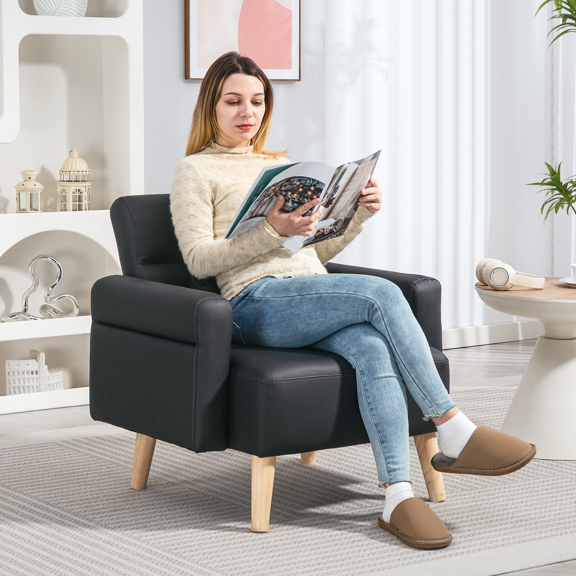 Mujer en jeans lee una revista, sentada en un sillón negro. Patas de madera, suéter beige y zapatillas marrones.