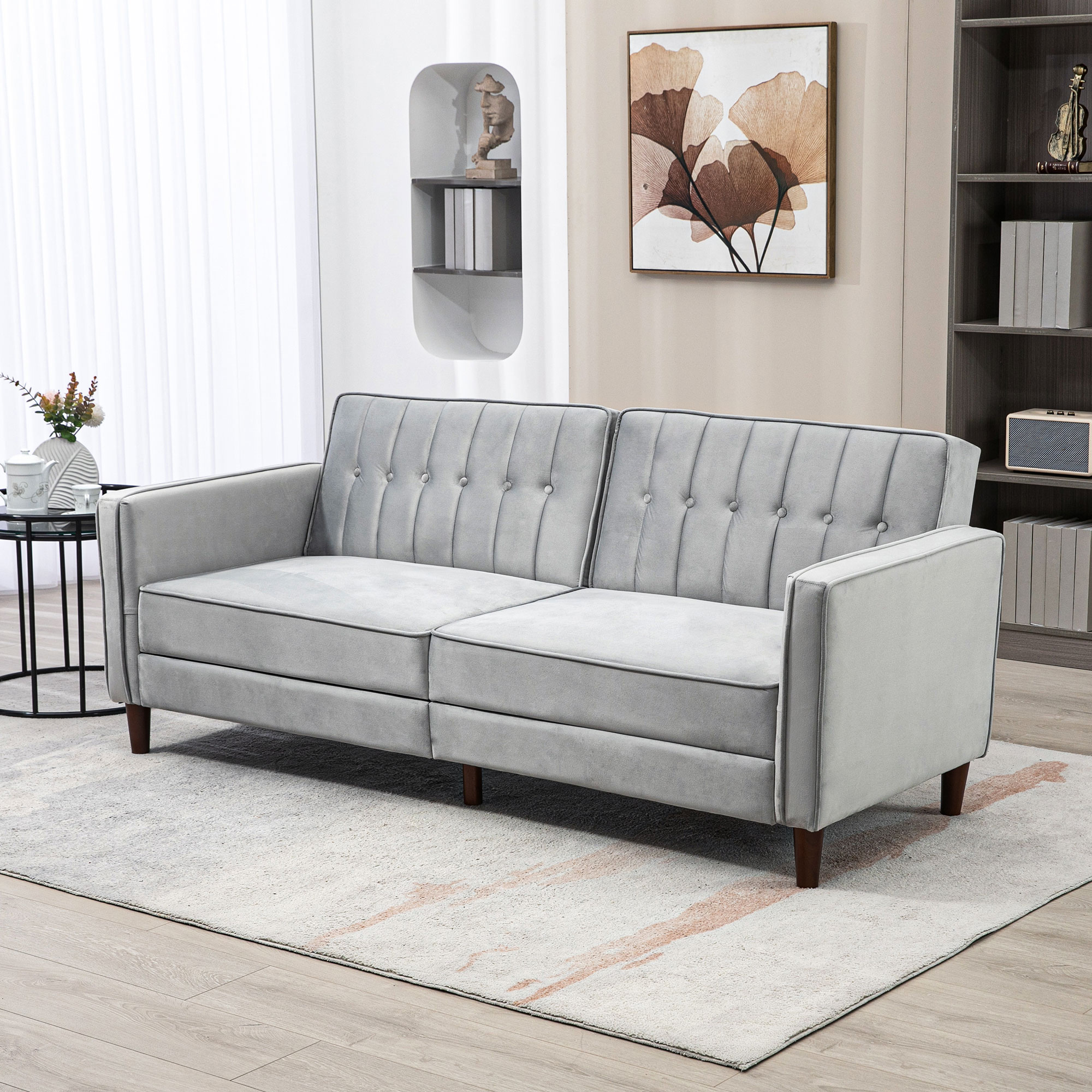 Un sofá gris en una sala de estar. Hay una pintura, estanterías y una alfombra. Flores y una mesa están en la escena.