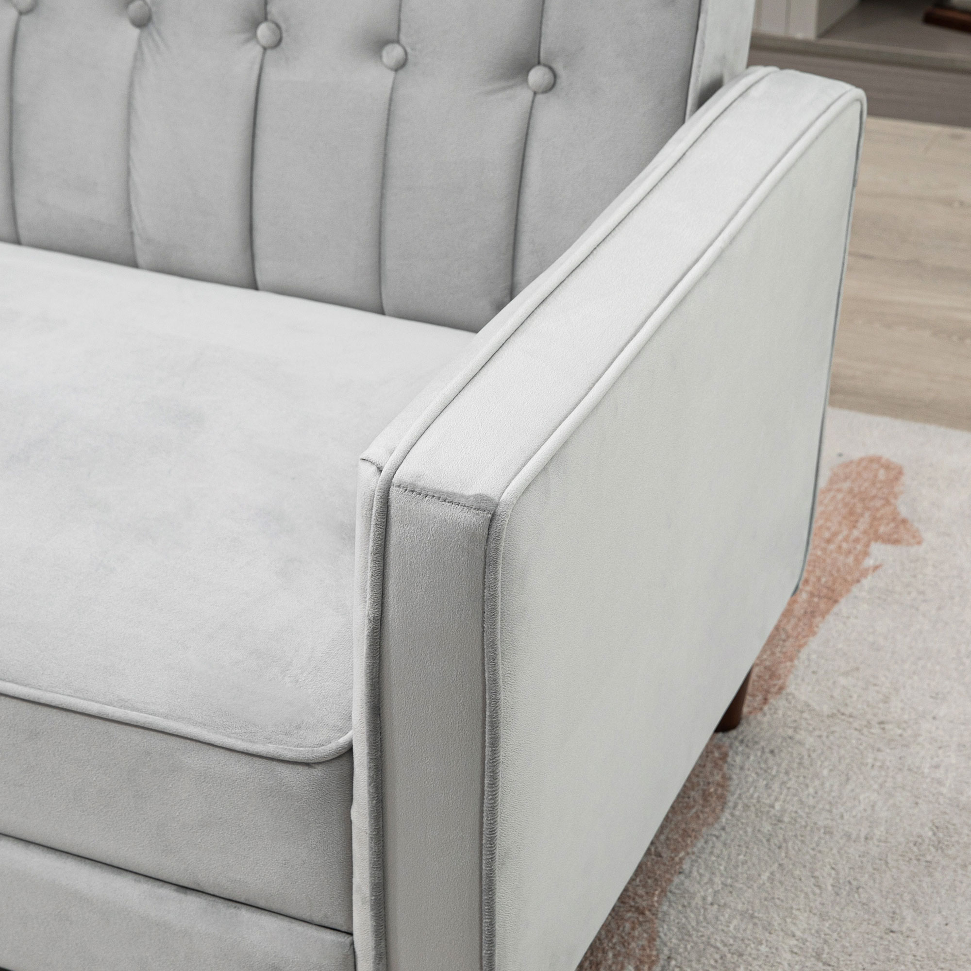 Un sofá gris claro con respaldo abotonado y reposabrazos está en una habitación. El sofá está sobre una alfombra estampada.