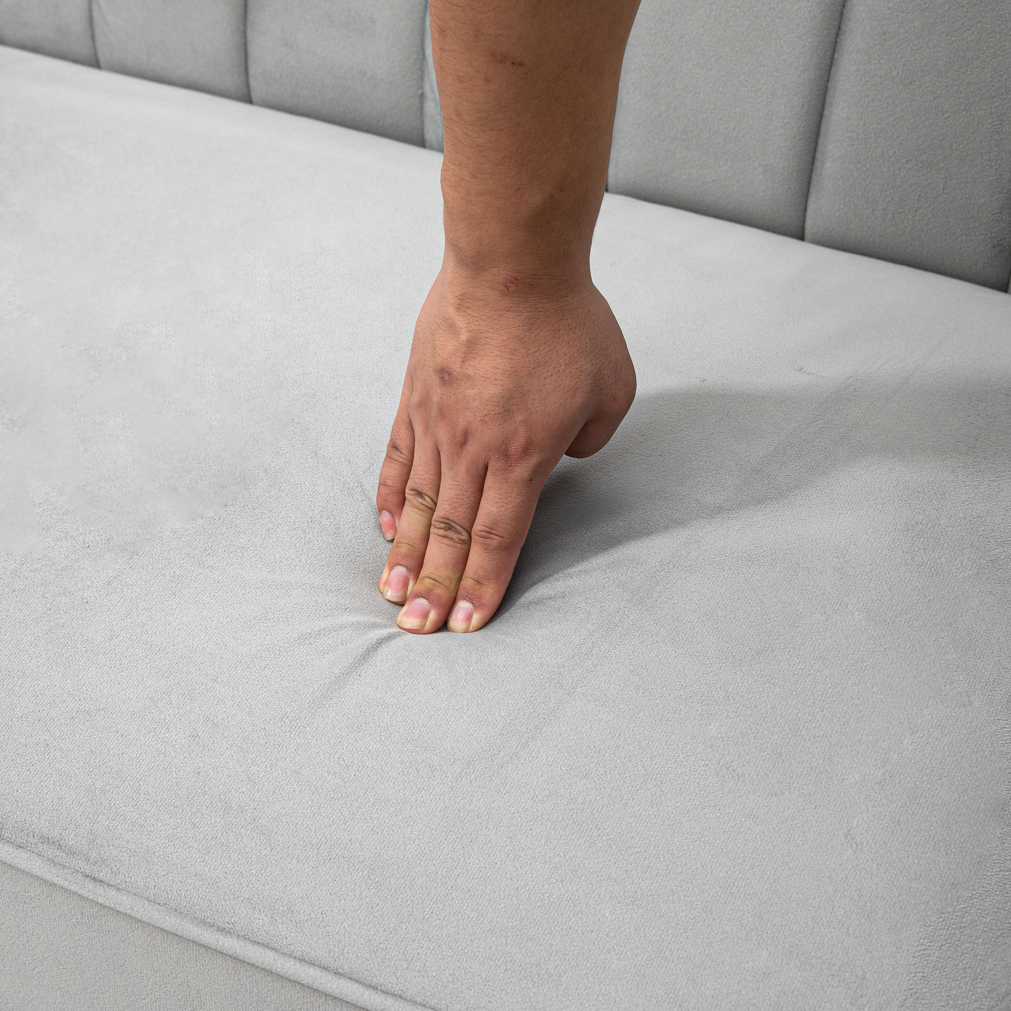 Una mano presiona sobre una superficie tapizada gris claro, posiblemente un sofá. Los dedos de la mano están extendidos.