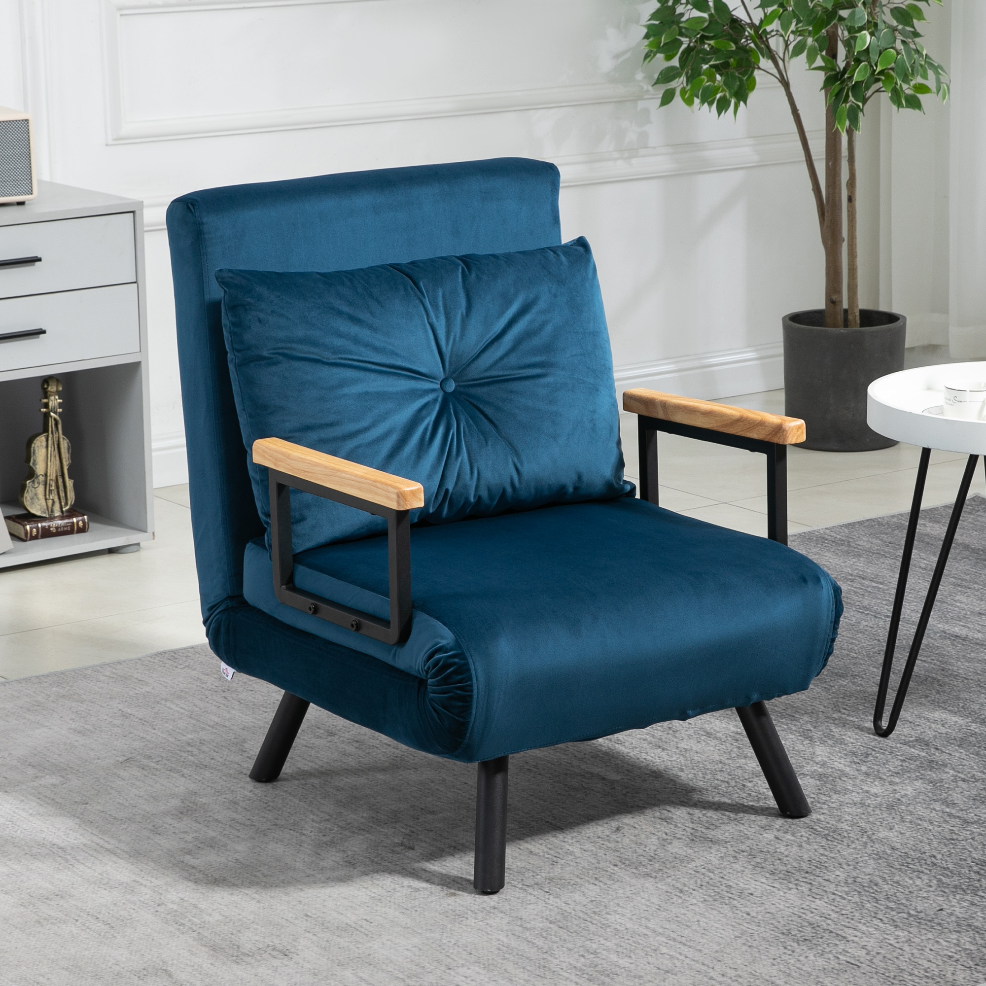 Un sillón color turquesa con reposabrazos de madera y un cojín abotonado, sobre una alfombra gris. Una planta y una mesa auxiliar al fondo.