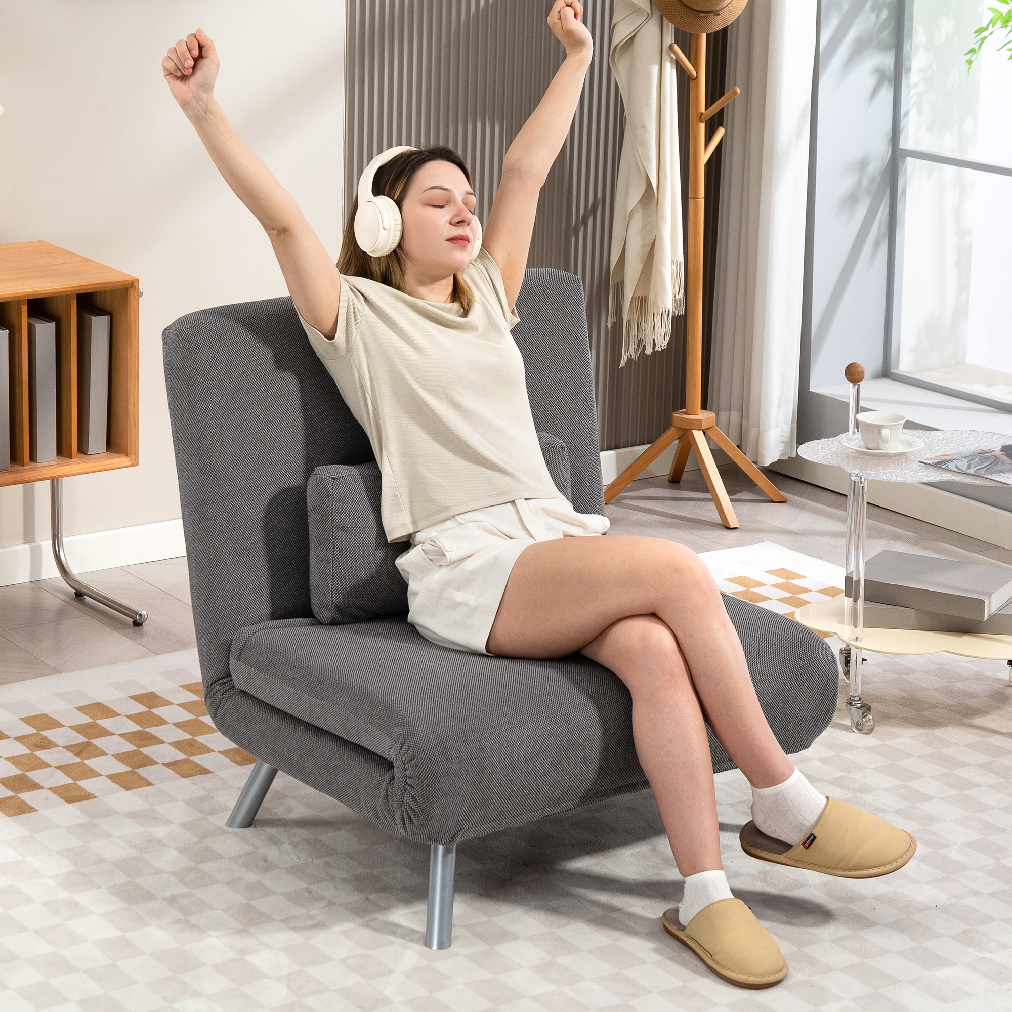 Una mujer sentada en una silla gris, con los brazos levantados, con auriculares. Lleva pantalones cortos y una camiseta. Hay un perchero y una mesa cerca.