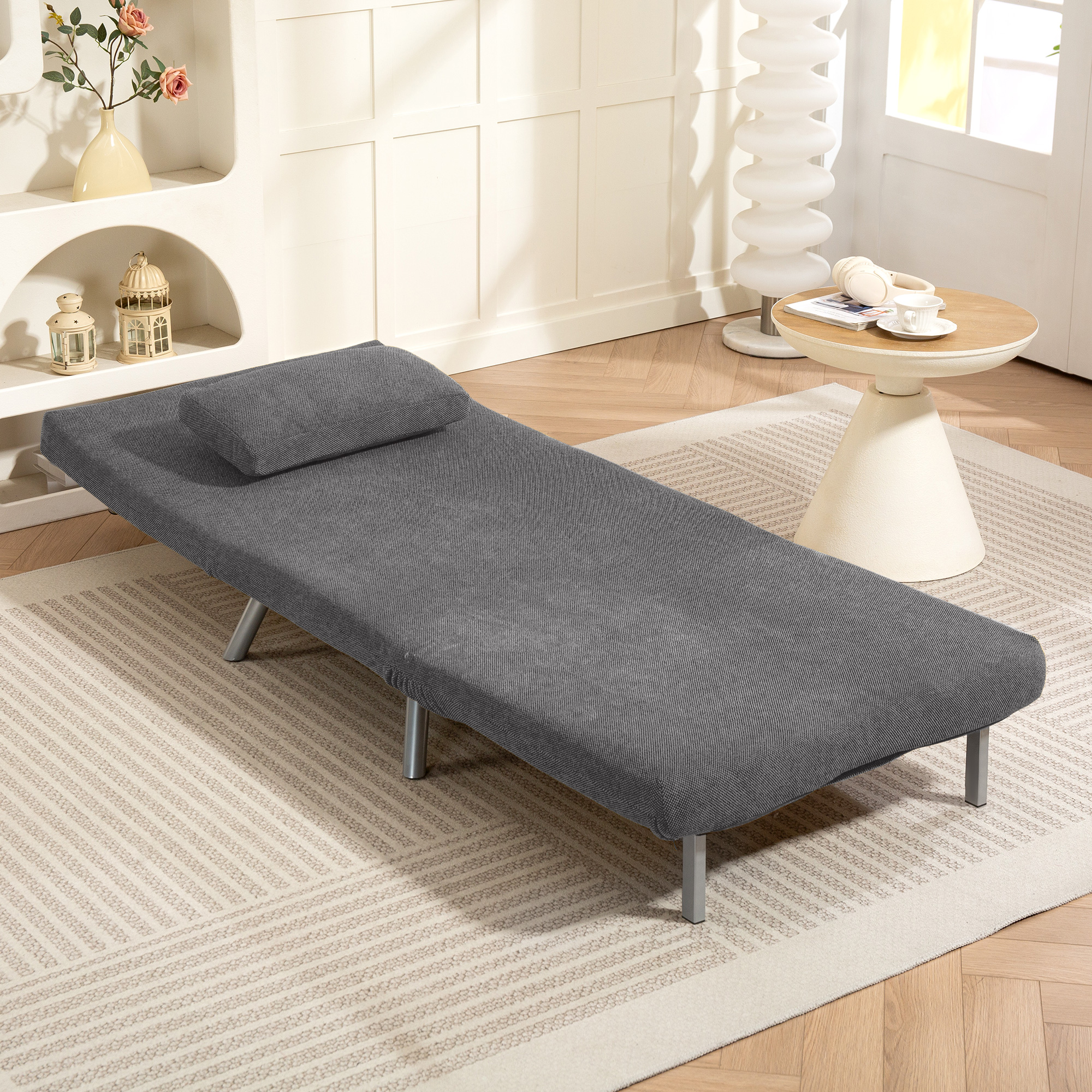 Una chaise longue gris con una almohada se encuentra sobre una alfombra estampada. Una mesita con una taza está al lado y un armario detrás.
