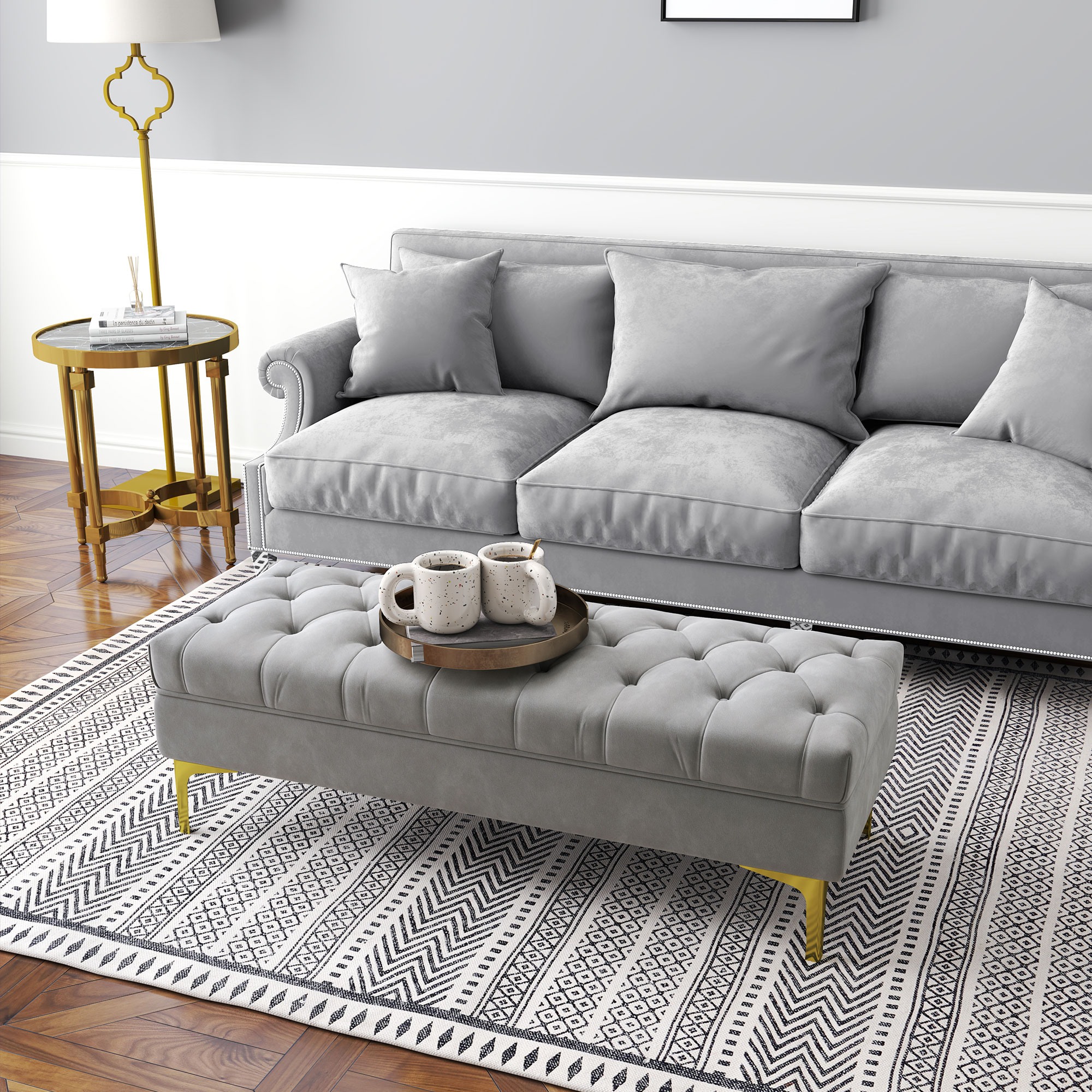 Salón con sofá gris, otomana y alfombra estampada. Dos tazas en una bandeja. Mesa auxiliar y lámpara doradas.