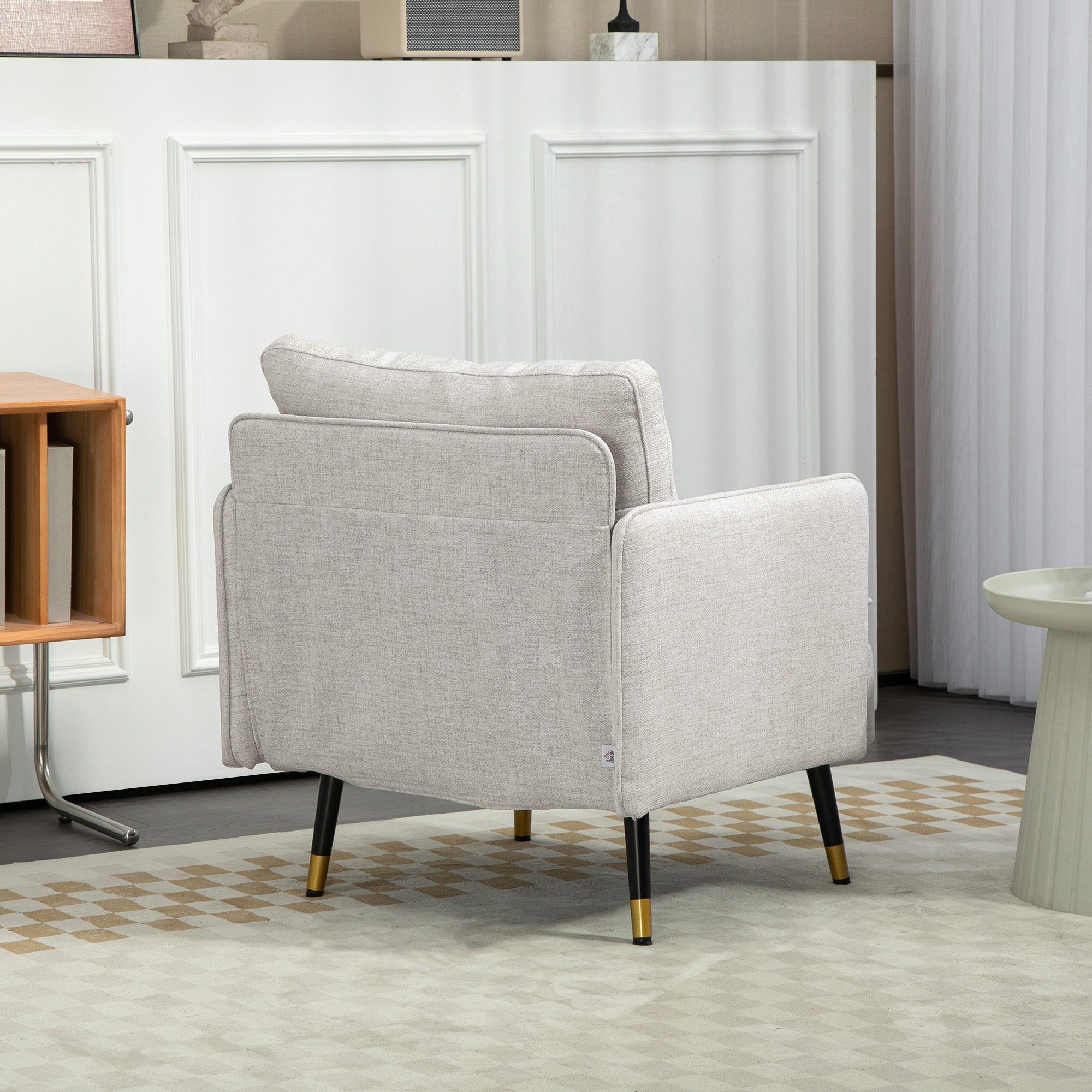 Un sillón beige con patas negras y puntas doradas, colocado sobre una alfombra estampada. A la izquierda, una estantería de madera.