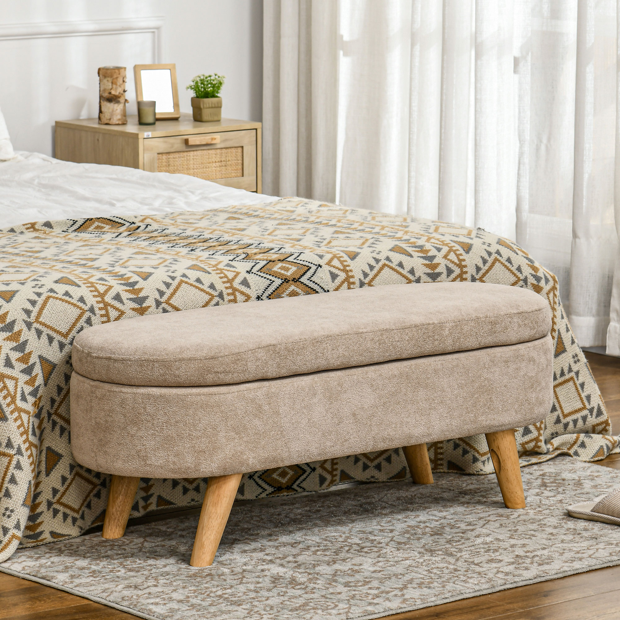 Un banco beige con patas de madera está frente a una cama. También se ven una mesita de noche y una manta estampada.