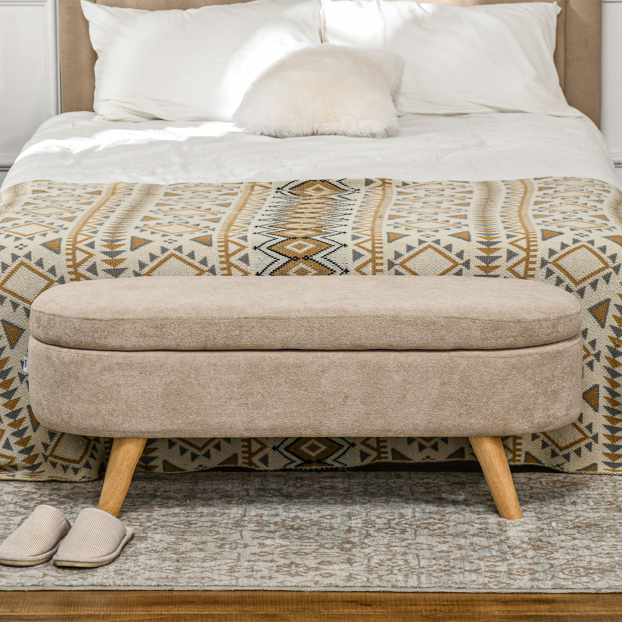 Una escena de dormitorio con una cama cubierta con sábanas blancas y una manta estampada. Un banco beige y zapatillas están al frente.
