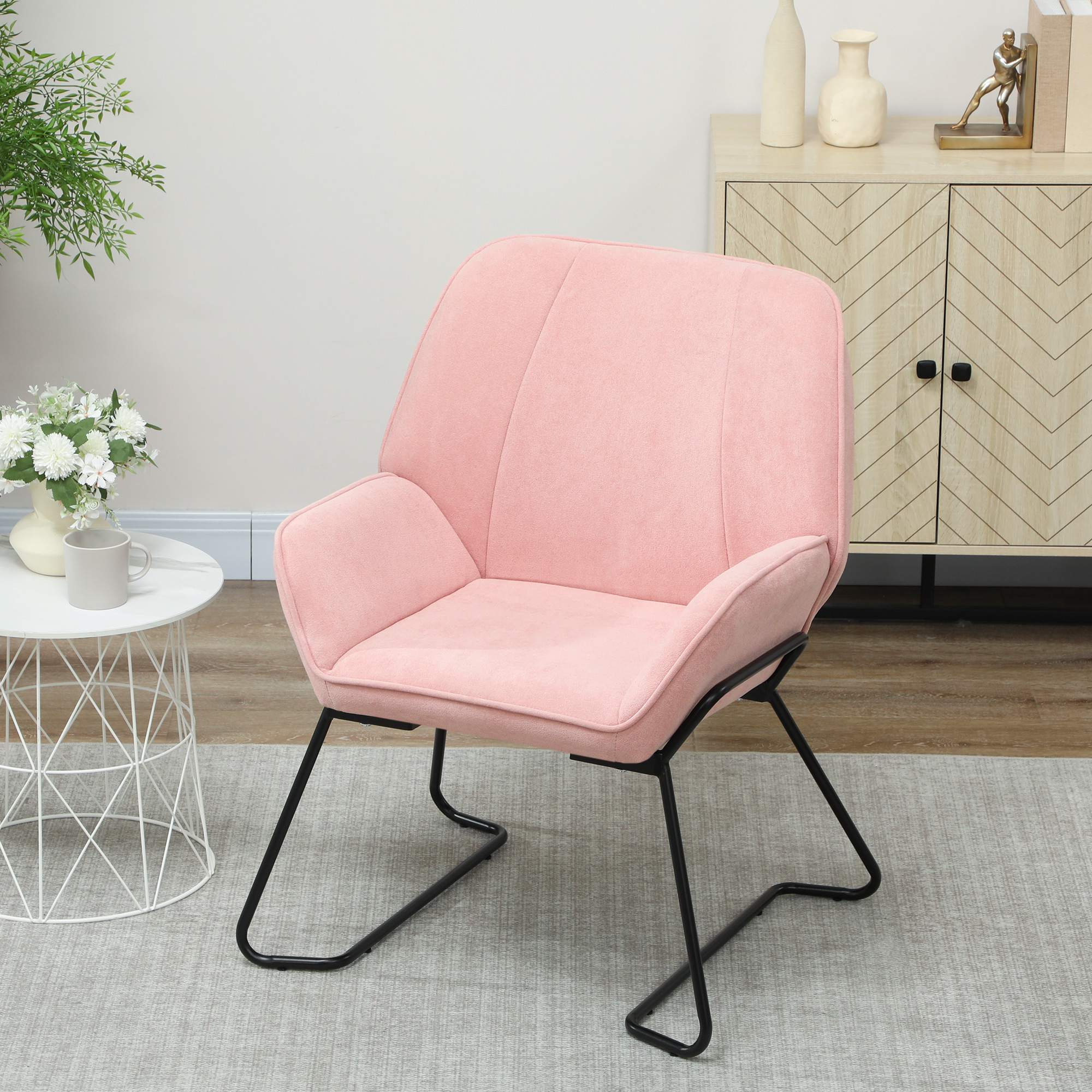 Sillón rosa con patas negras sobre una alfombra gris. Mesa auxiliar blanca, mueble y plantas en el fondo.