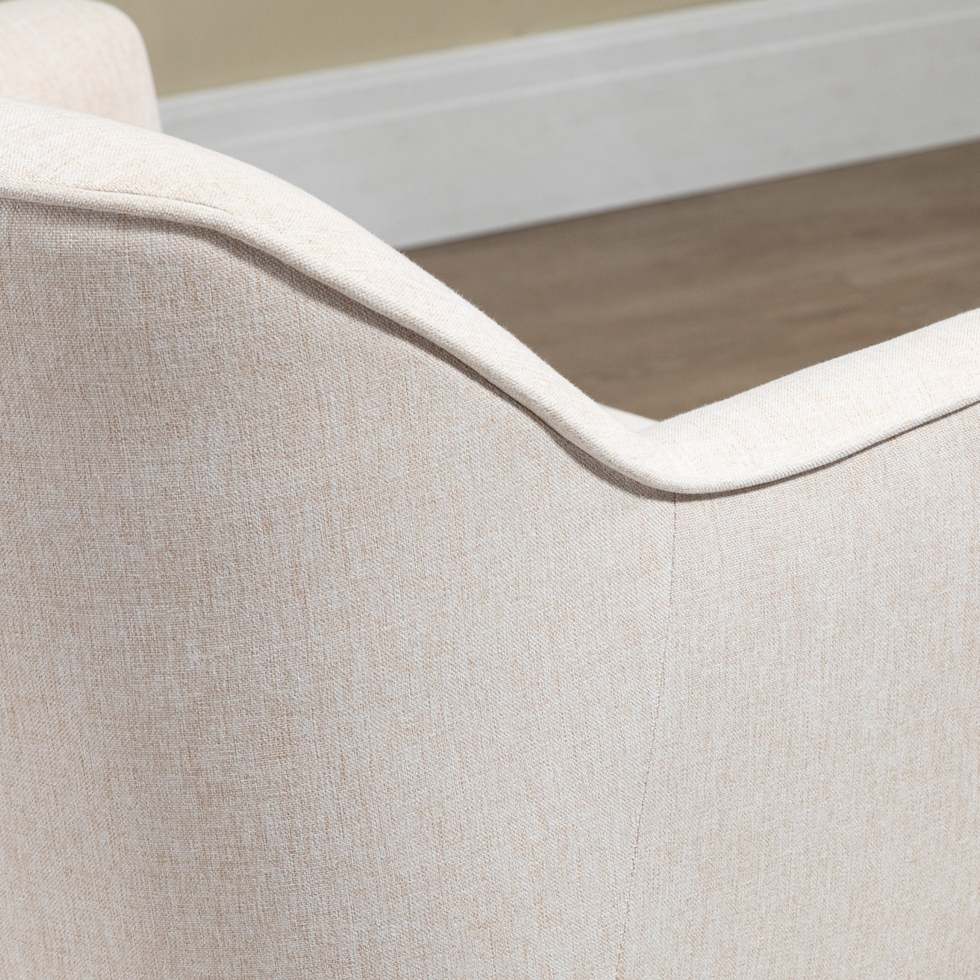 Primer plano del respaldo de una silla de tela beige. Se ve la textura con un diseño curvo. El fondo muestra una pared y un suelo.