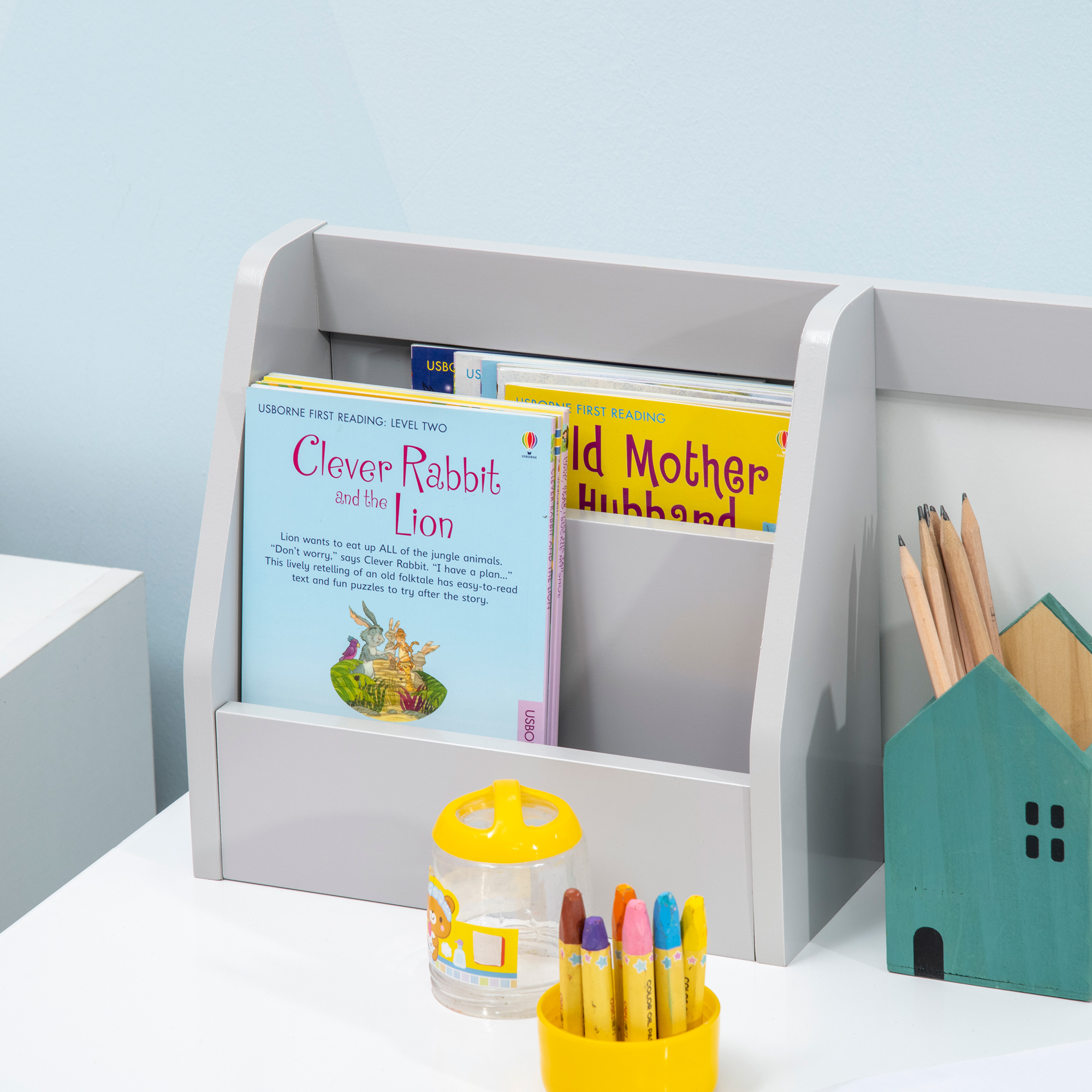 Una estantería gris con libros infantiles, crayones, lápices y recipientes en forma de casa se encuentra sobre una mesa blanca.