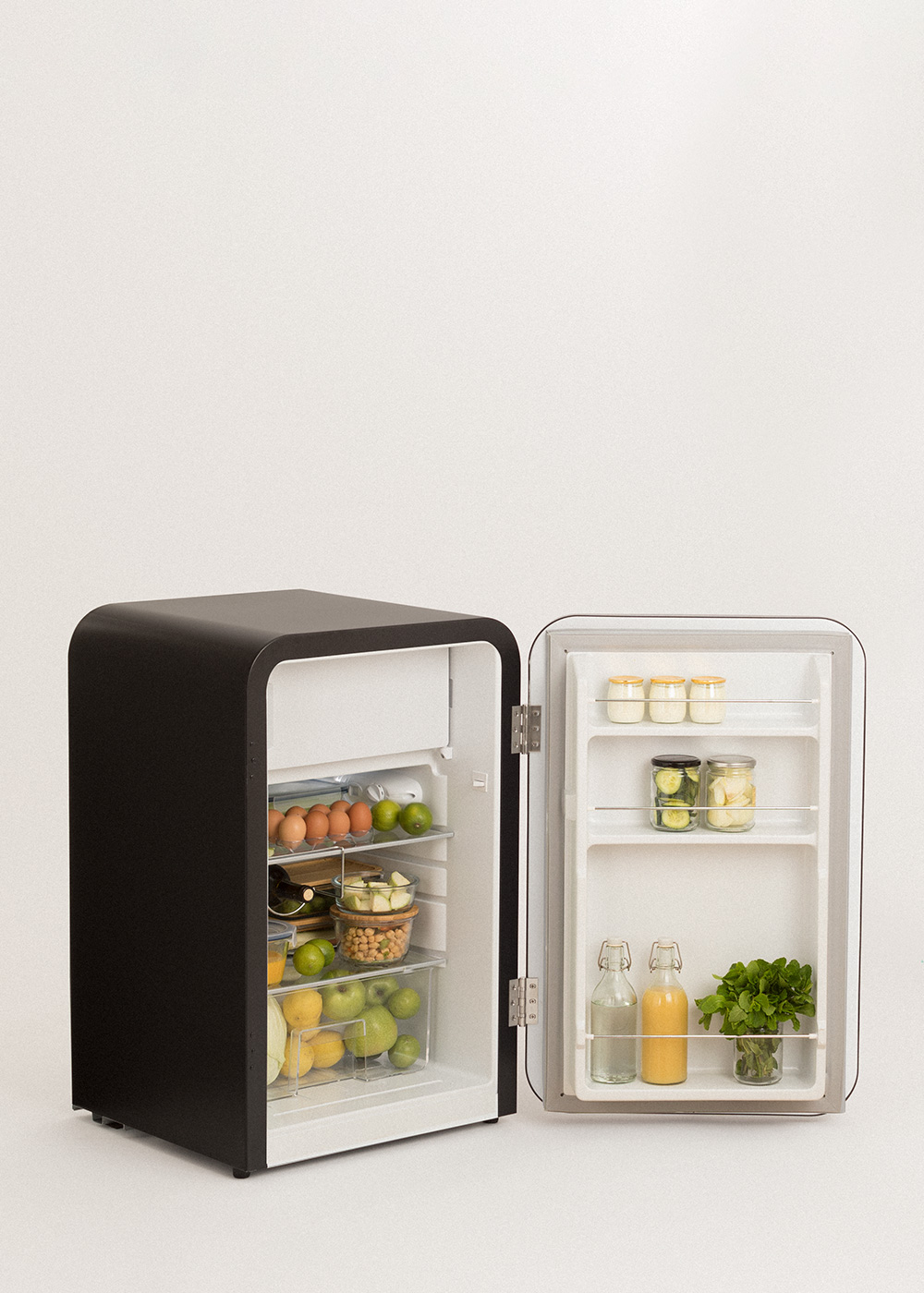 Una mini nevera negra está abierta, mostrando alimentos como huevos, frutas, frascos con artículos y botellas. El fondo es blanco.