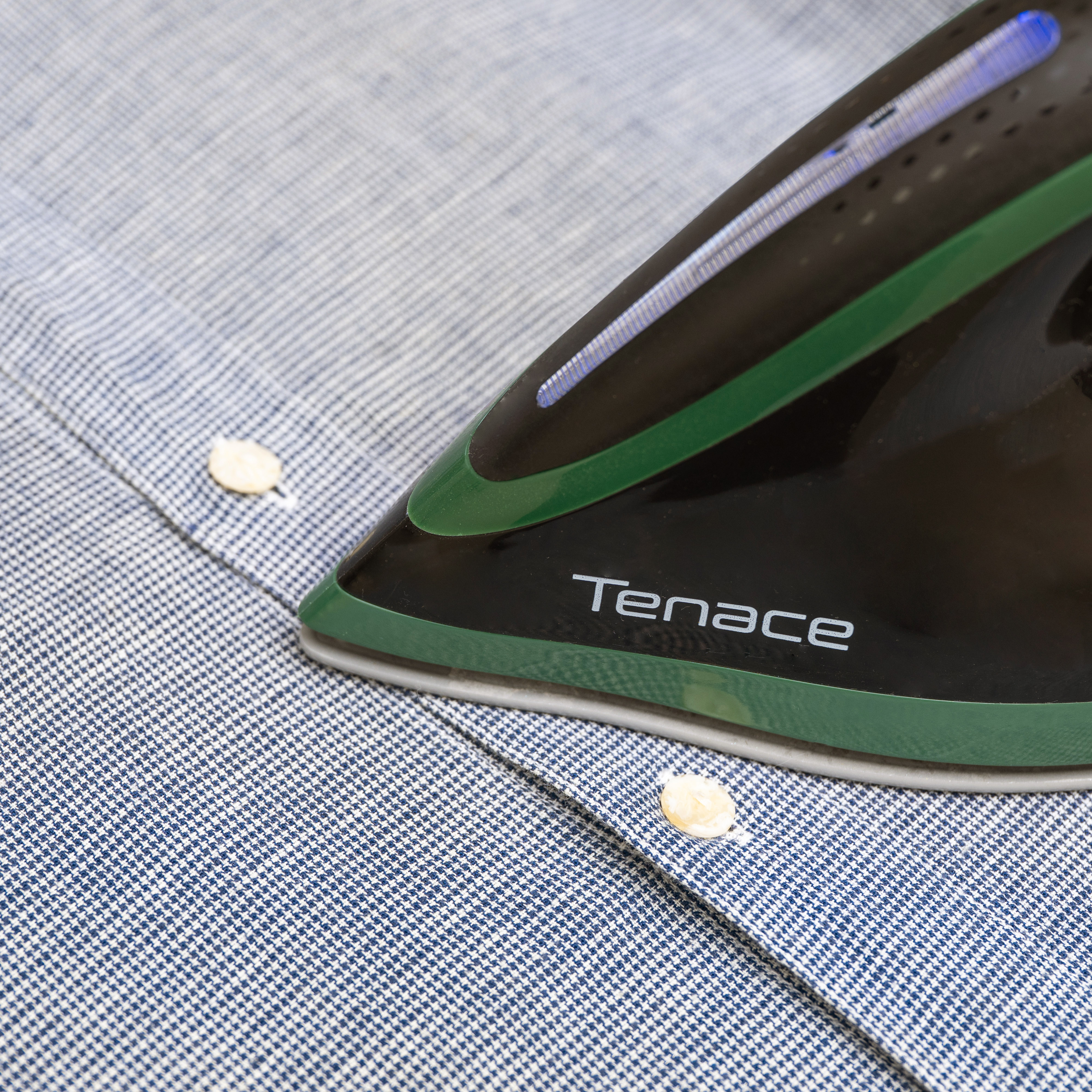 Una plancha negra y verde está sobre una camisa a cuadros azul y blanco. Se ven botones. La plancha dice 'Tenace'.