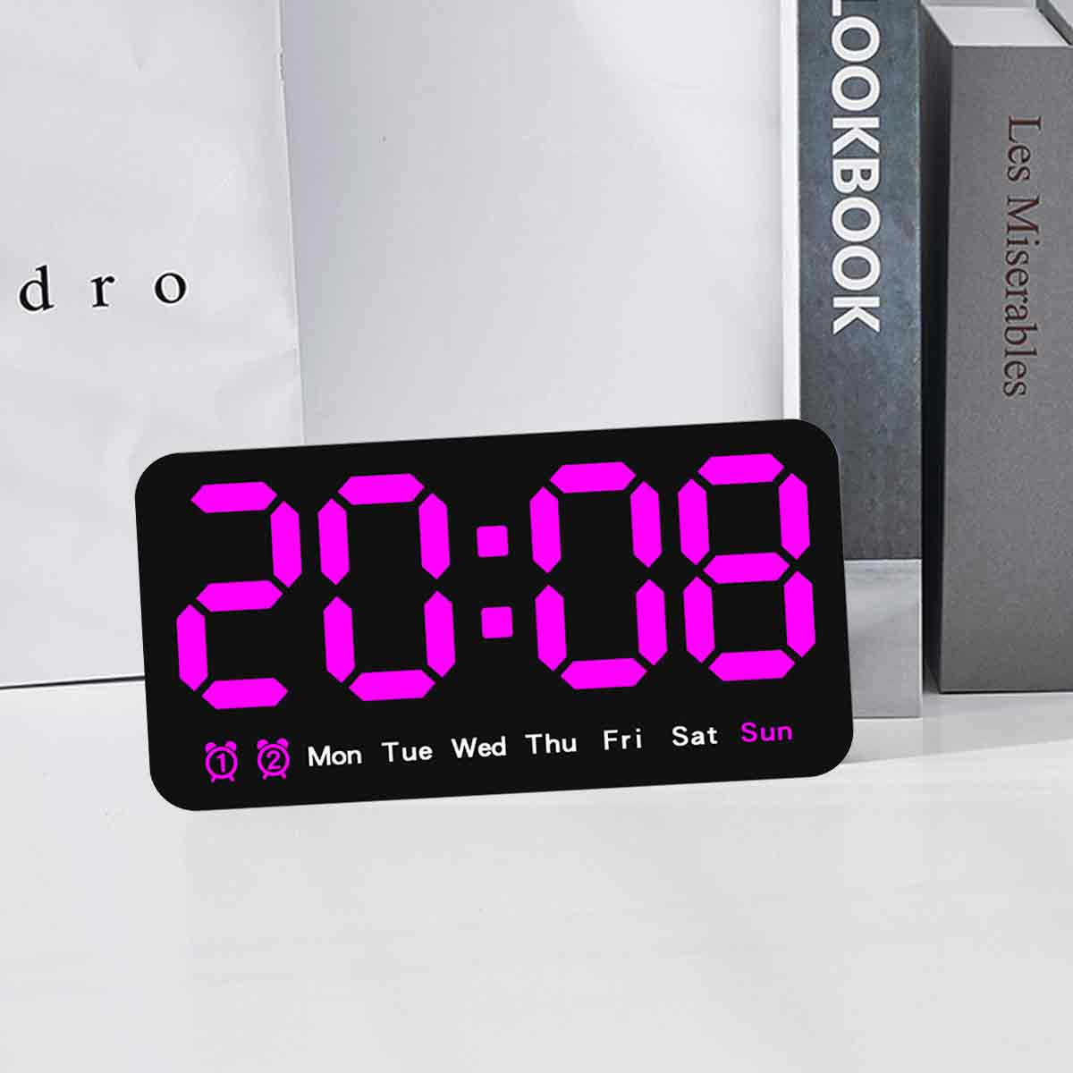 Eine Digitaluhr zeigt 20:08 in hellem Pink an. Im Hintergrund sind Bücher zu sehen. Die Wochentage sind unten.