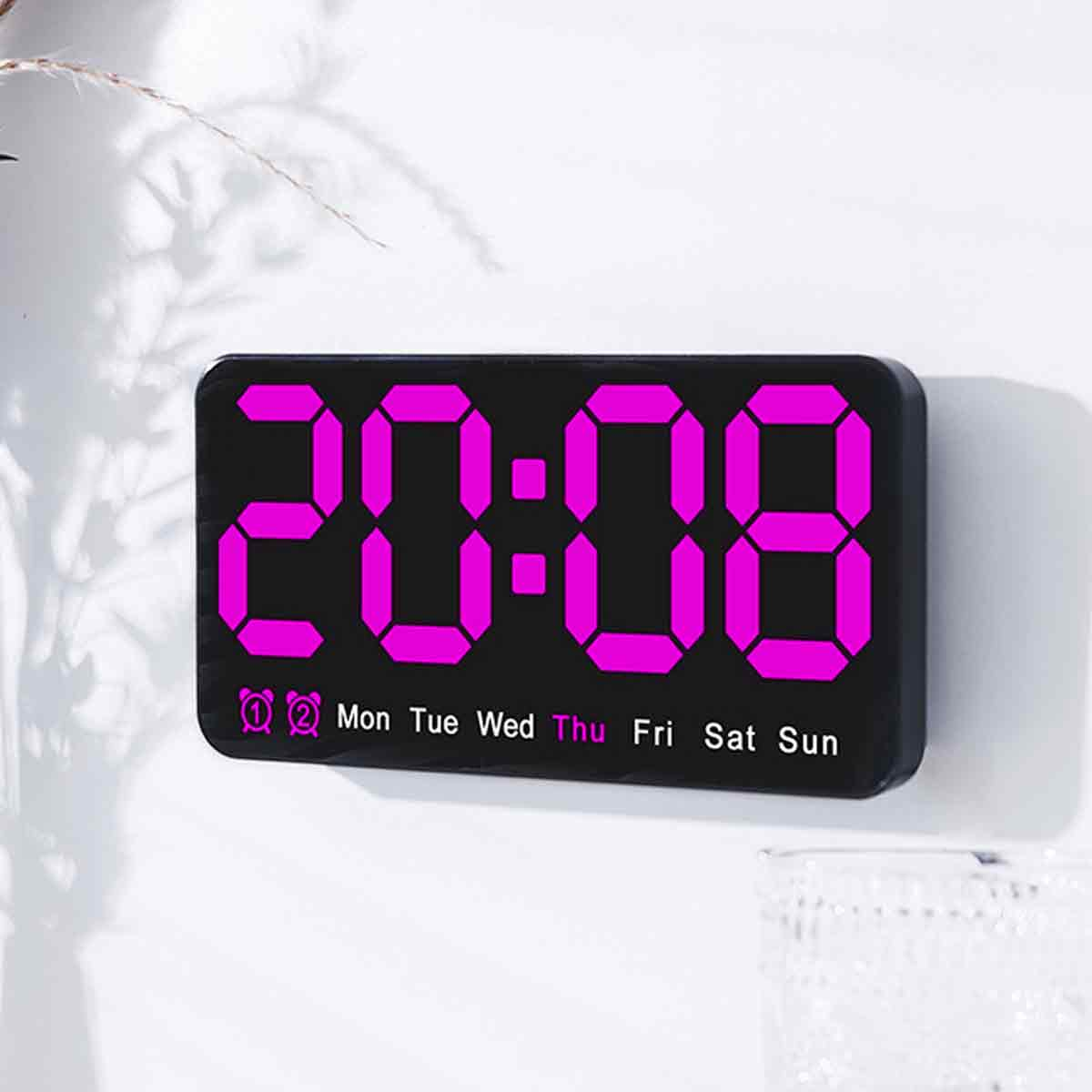 Eine Digitaluhr zeigt 20:08 in Pink an. Wochentage darunter. Die Uhr befindet sich auf einer weißen Oberfläche.