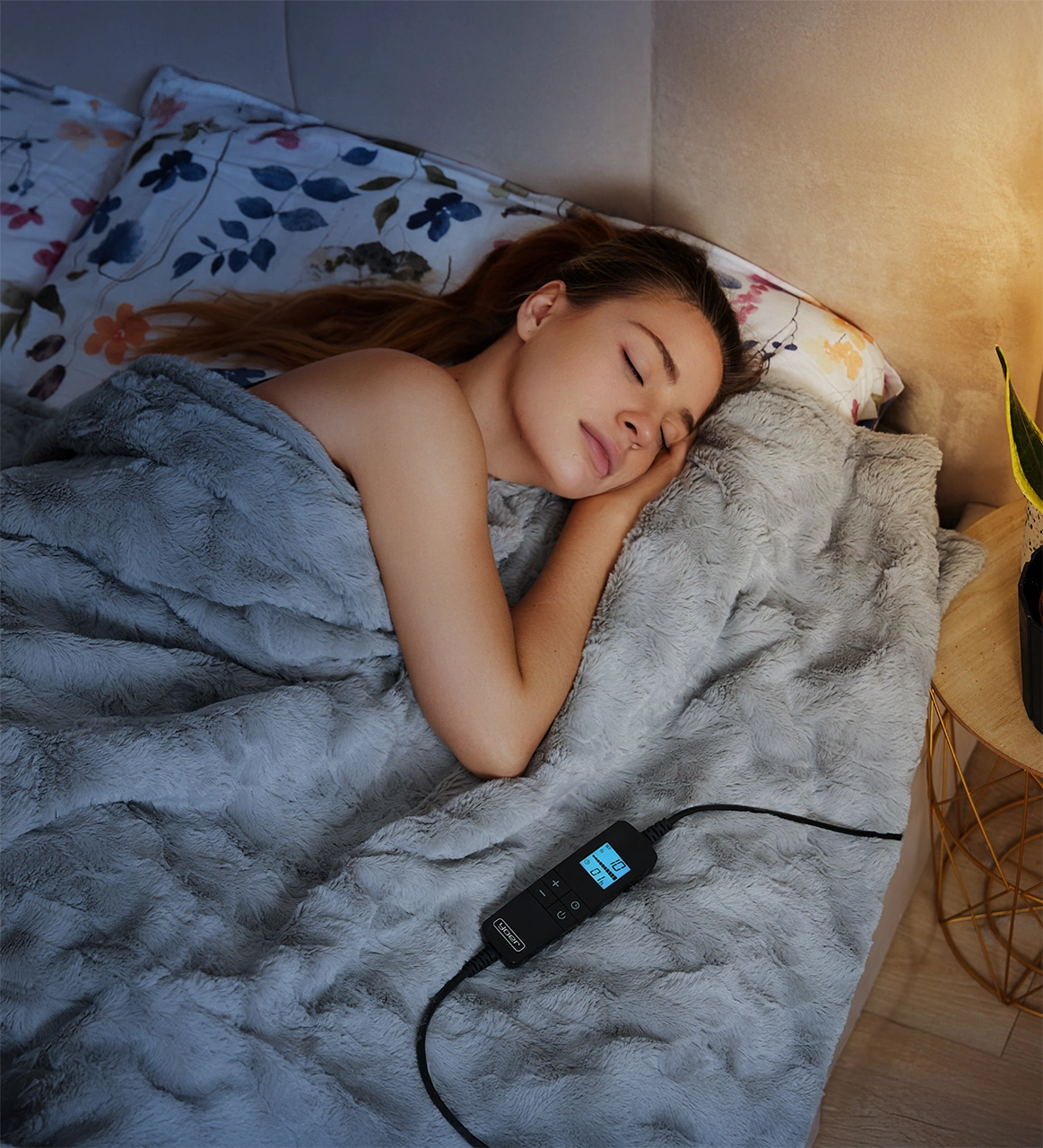 Kobieta śpi pod szarą kołdrą na łóżku. Widoczny panel sterowania z wyświetlaczem. Kwiatowe poduszki w tle.
