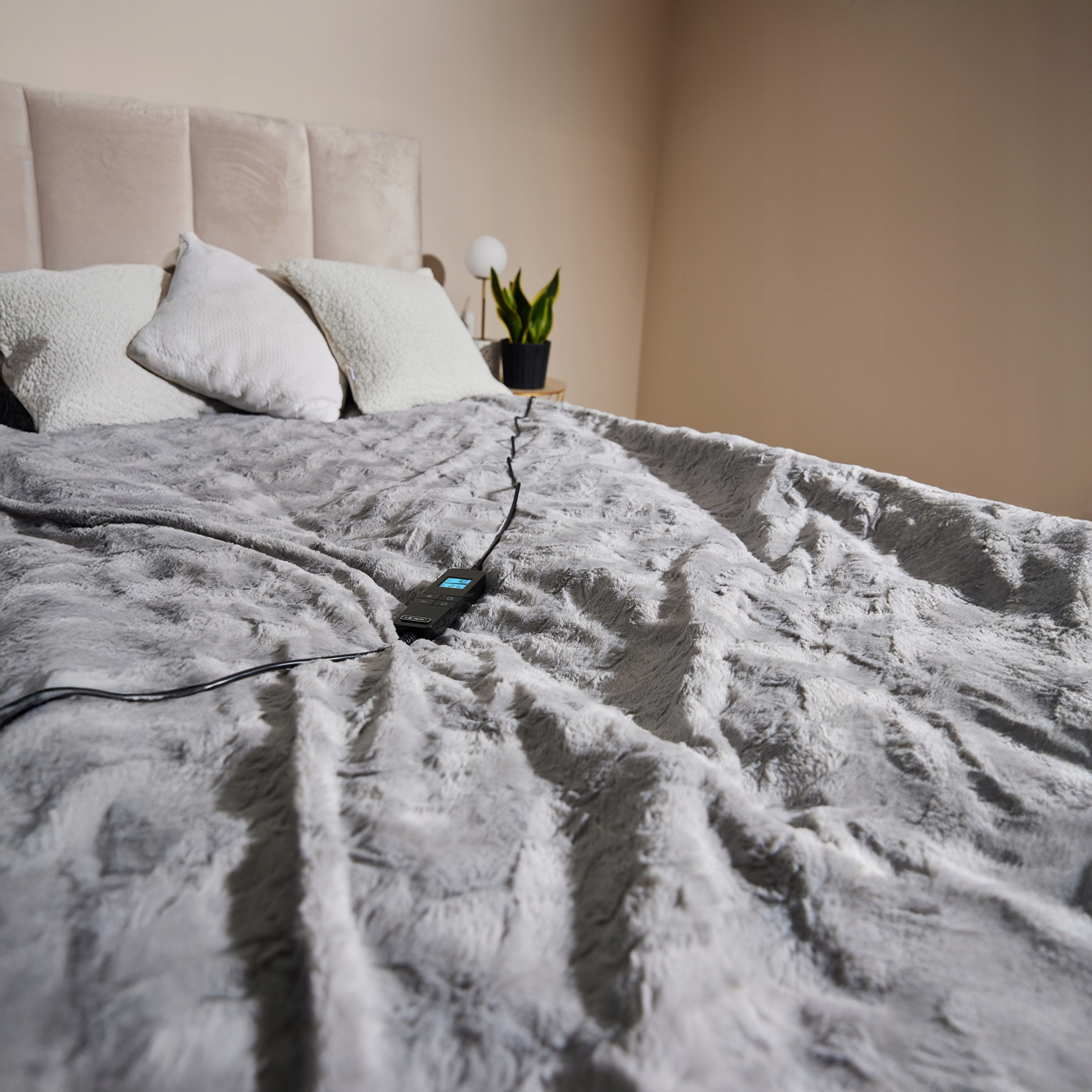 Łóżko z szarym kocem i poduszkami. Kontroler jest na kocu. Roślina na stoliku nocnym.