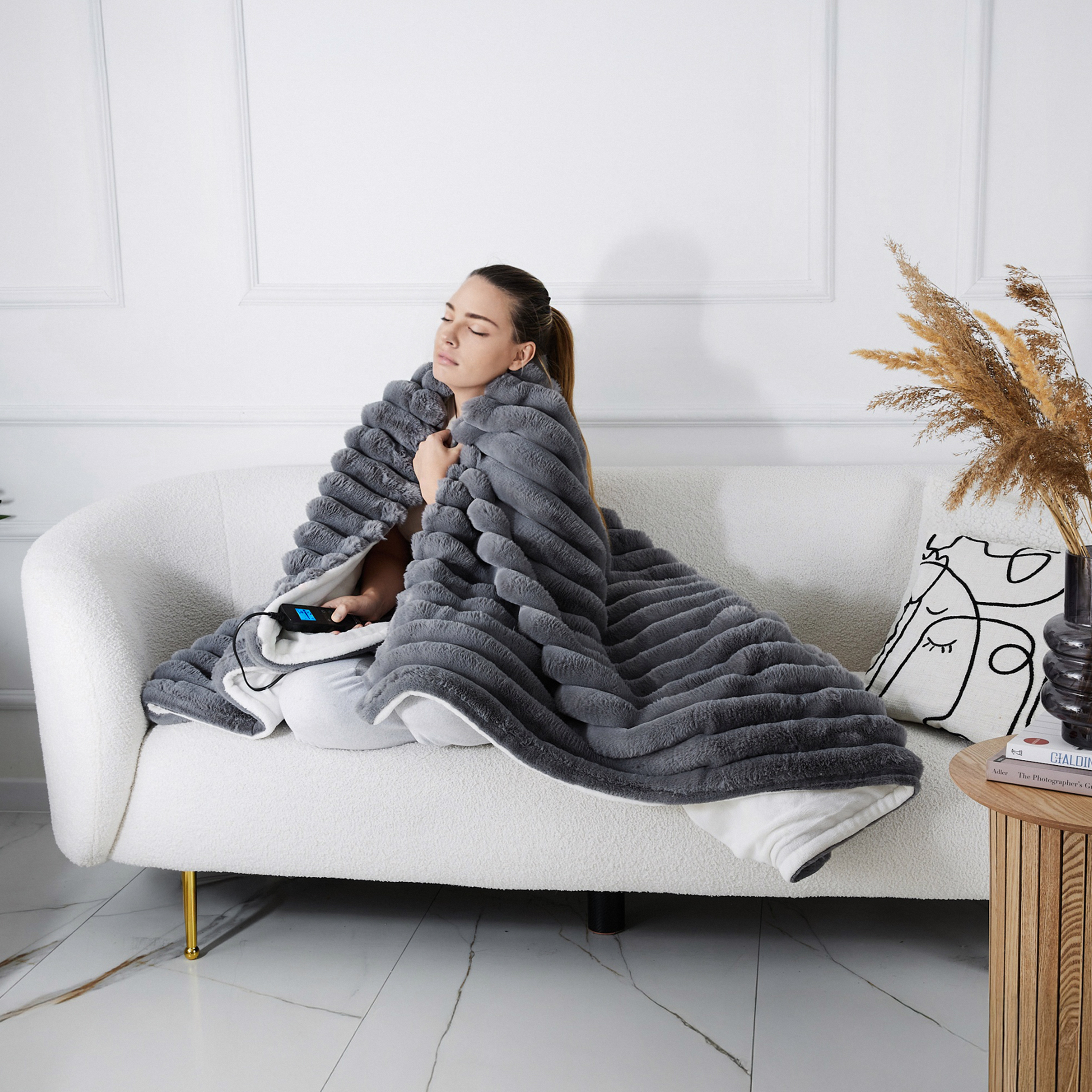 Kobieta siedzi na białej sofie, owinięta szarym kocem. Stolik boczny ma książkę i wazon.