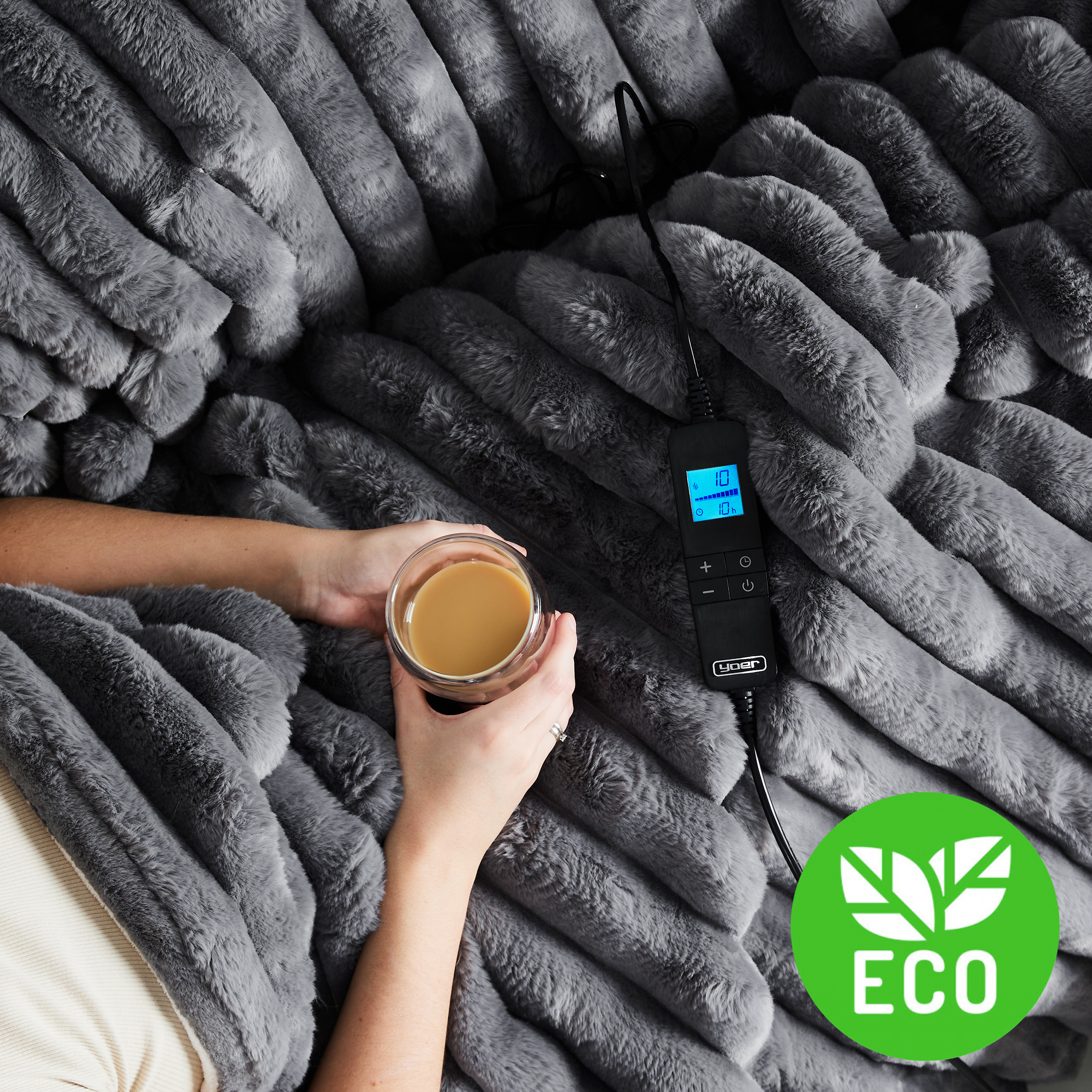 Osoba trzyma kawę w szklance na szarym kocu z imitacji futra. Widoczne jest ekologiczne logo.