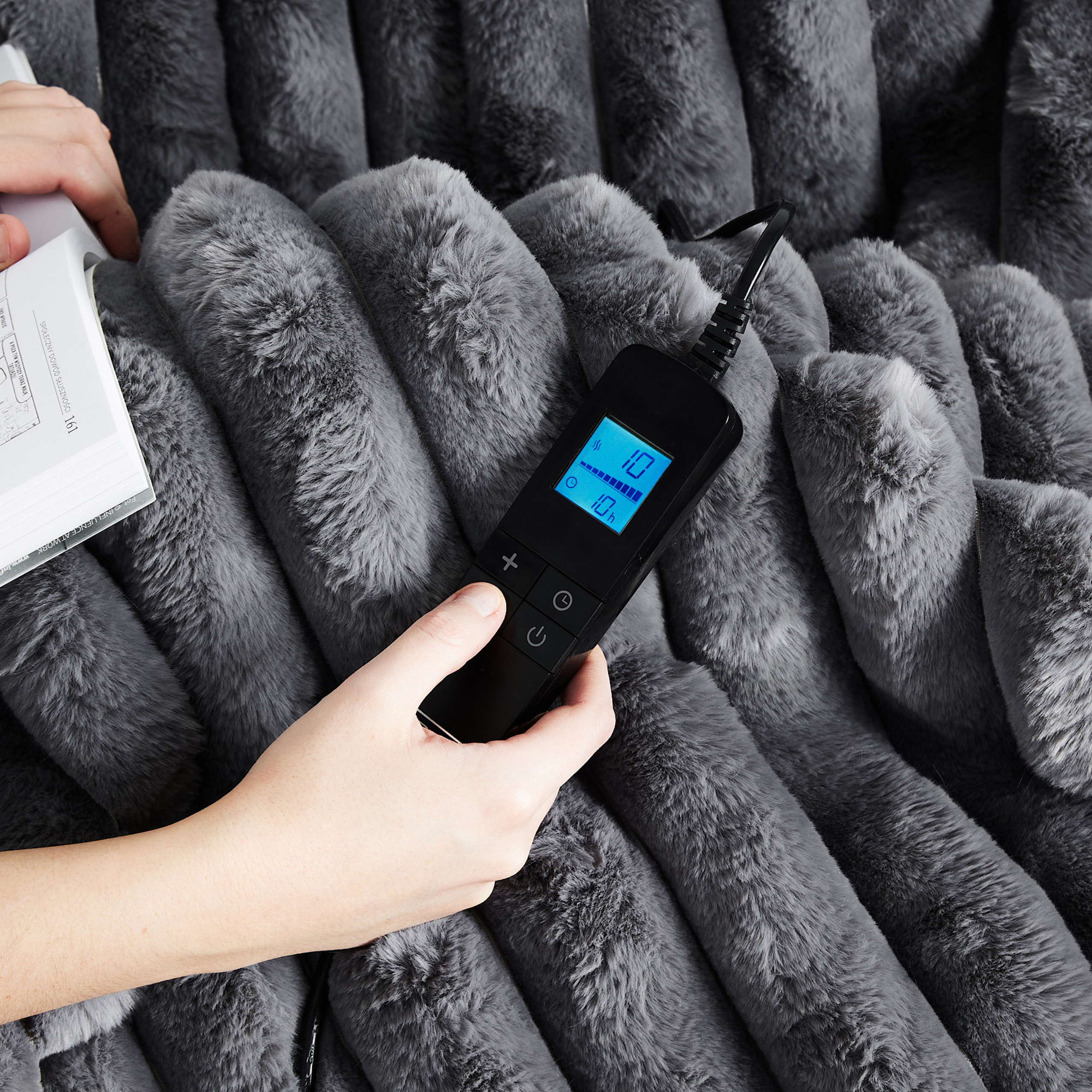 Ręka trzyma pilota i książkę nad szarą, puszystą kołdrą. Pilot wyświetla ustawienia.