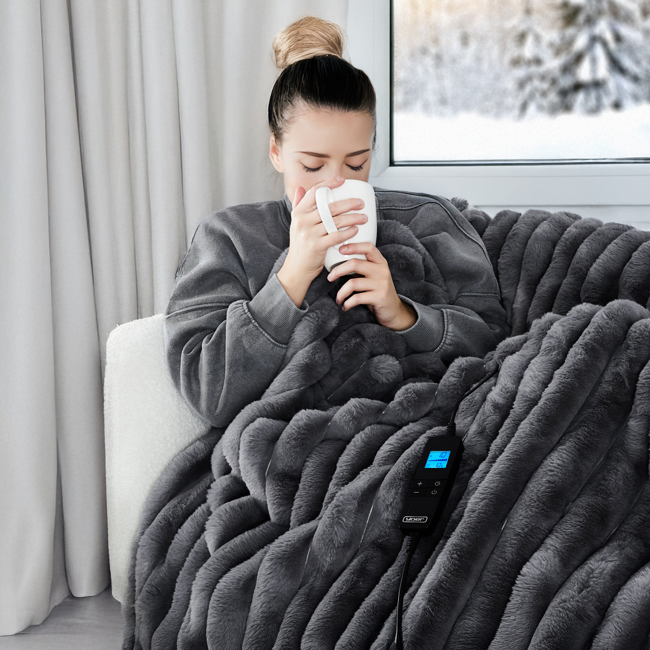 Kobieta w szarej bluzie otulona szarym kocem, pije z białego kubka przy oknie.