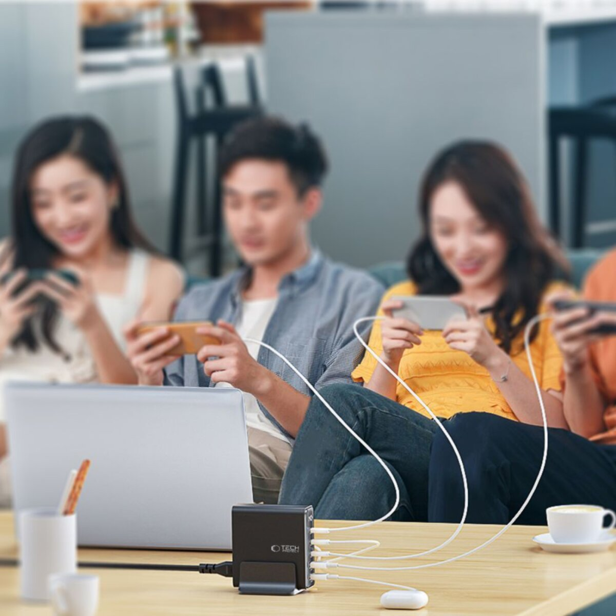 Des personnes utilisent des téléphones, connectés à un chargeur noir sur une table. Un ordinateur portable et une tasse sont également présents.