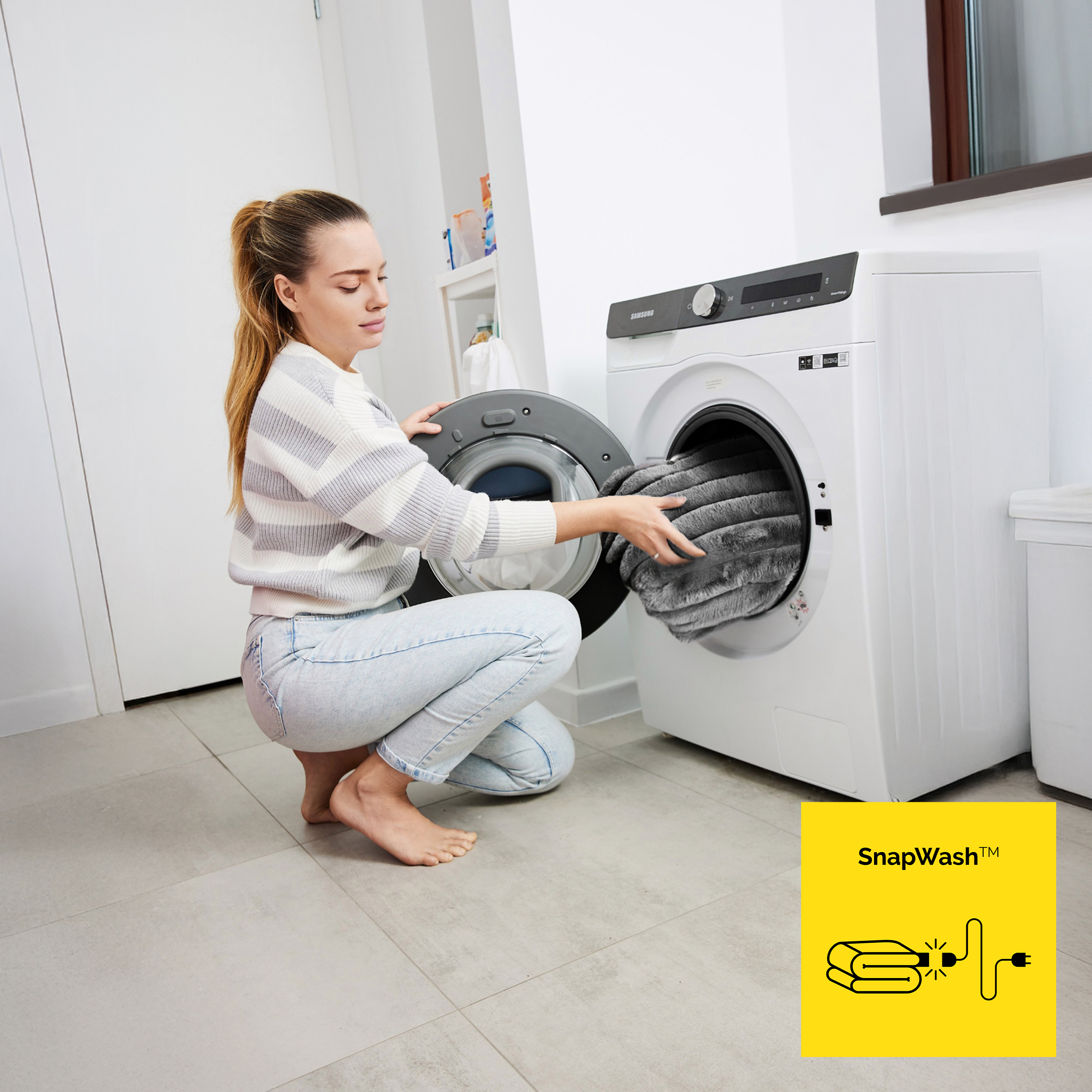 Kobieta wkłada szary koc do białej pralki. Ma na sobie koszulę w paski i dżinsy.