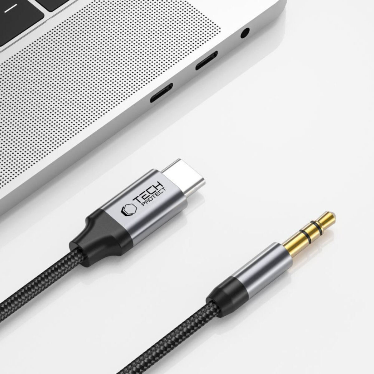 A USB-C to 3.5mm audio cable on a white surface next to a laptop. The cable has a metallic connector and black braid.