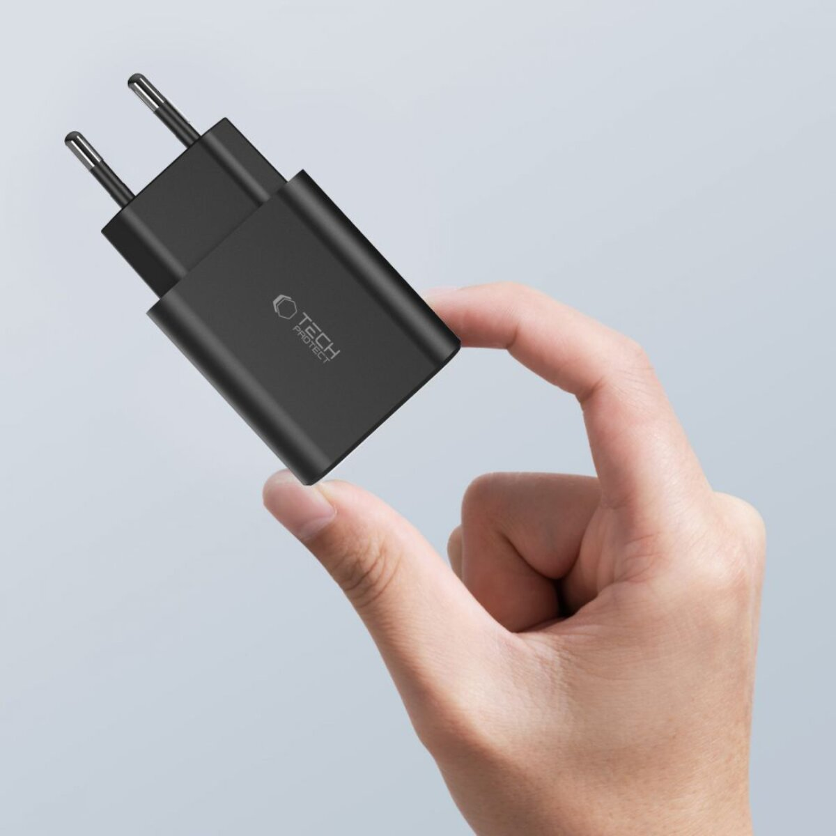 Een hand die een zwarte Tech Protect oplader vasthoudt. De oplader heeft twee pinnen en een logo. De achtergrond is lichtgrijs.