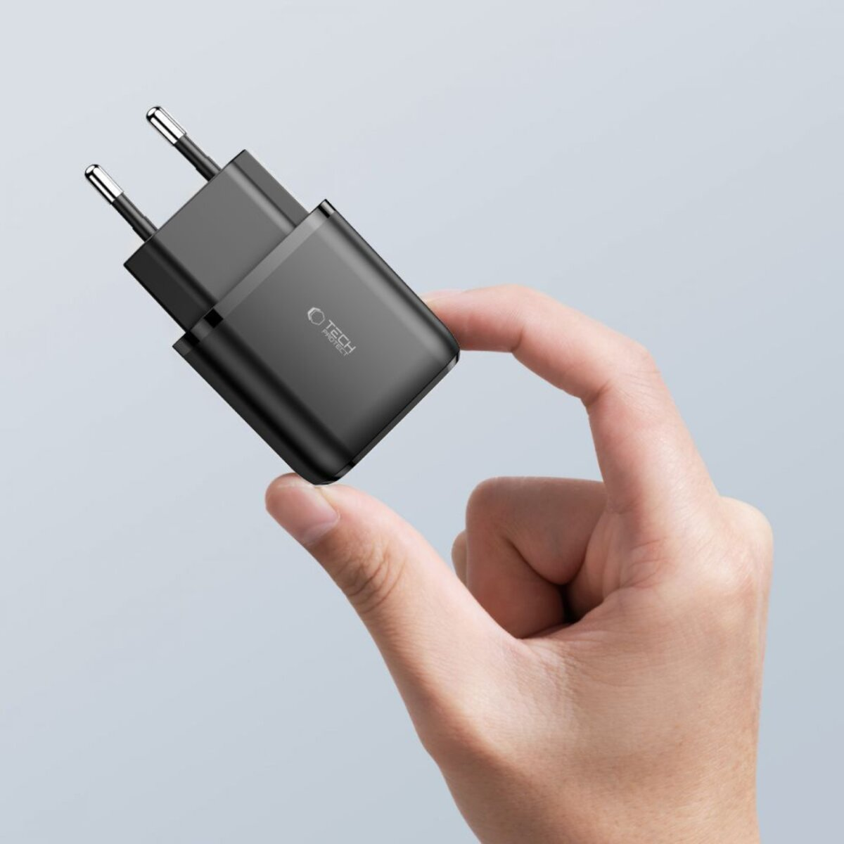 Une main tient un chargeur noir, avec deux broches argentées. Le chargeur porte l'inscription TECH PROTECT. Le fond est gris clair.