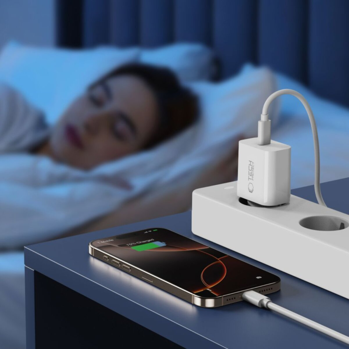 Ein Handy wird auf einem Nachttisch geladen, im Hintergrund schläft eine Person. Ein weißes Ladegerät ist eingesteckt.