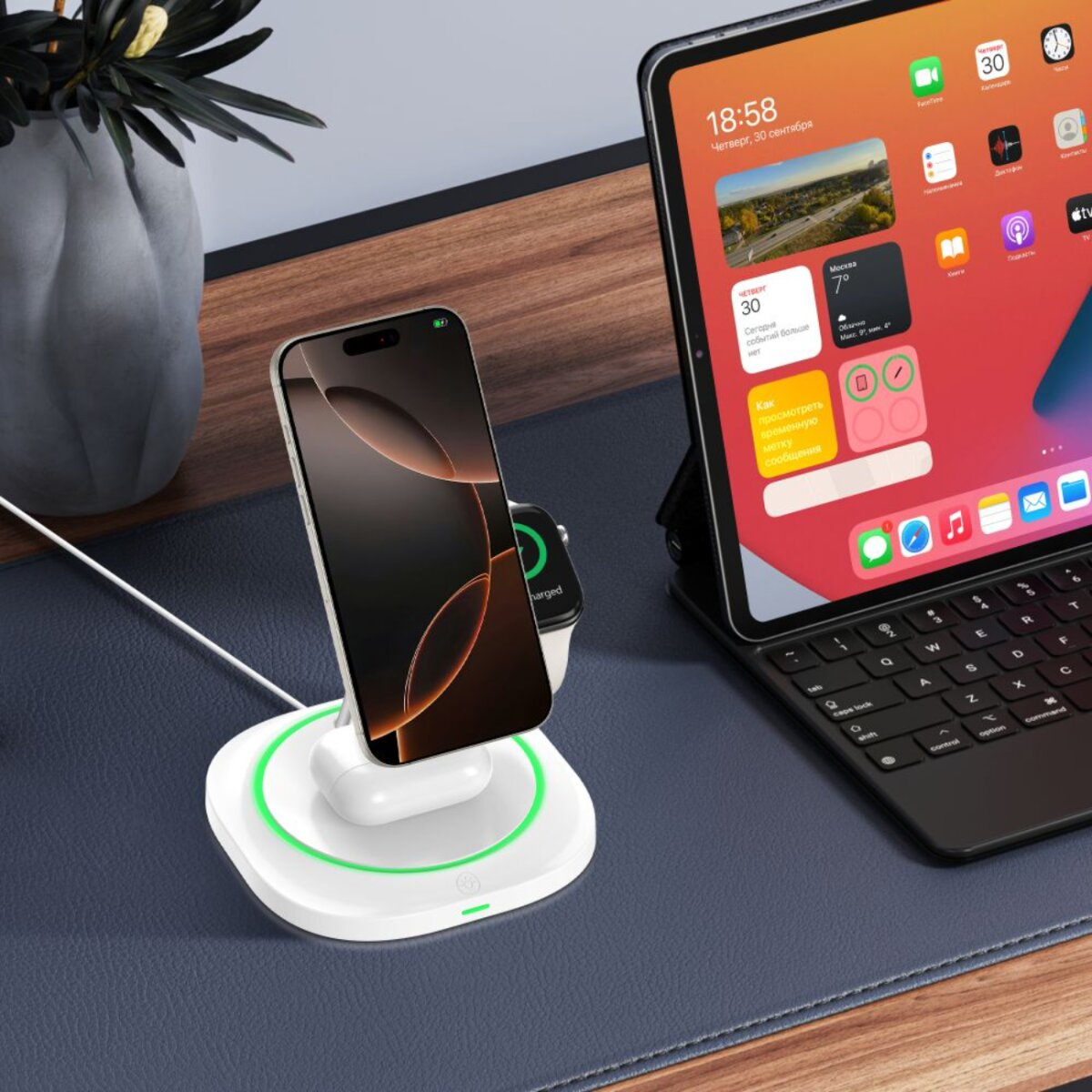 Ein Telefon, Ohrhörer und eine Smartwatch werden auf einem weißen Pad aufgeladen. Daneben steht ein iPad mit Tastatur, im Hintergrund eine Pflanze.