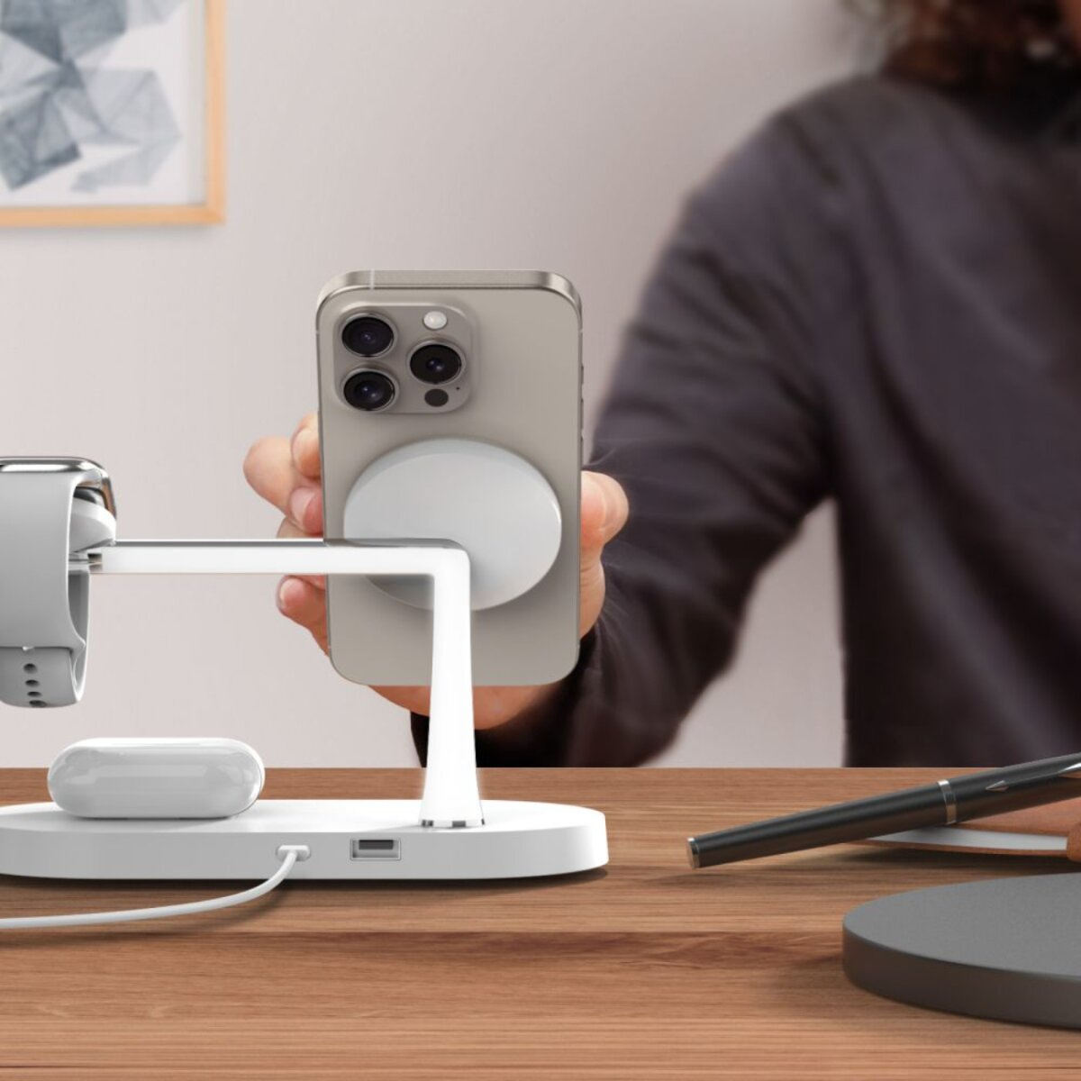 Une personne tient un téléphone au-dessus d'une station de charge avec une montre, des écouteurs et un stylo sur un bureau en bois.