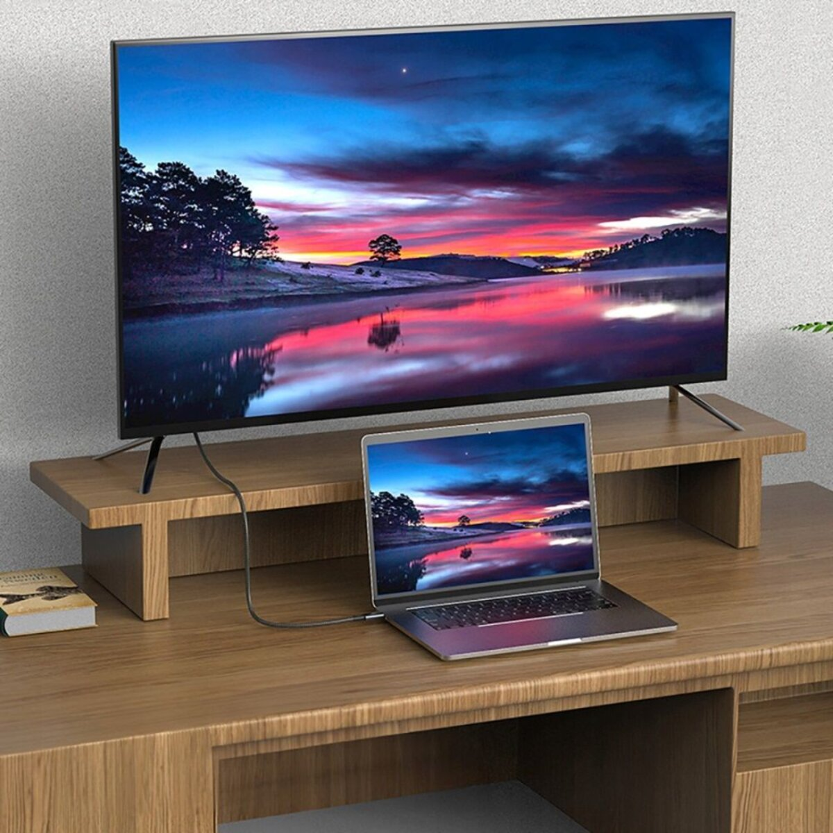 Un ordinateur portable sur un bureau en bois avec un support TV. La télévision affiche un paysage de coucher de soleil réfléchi dans un lac.
