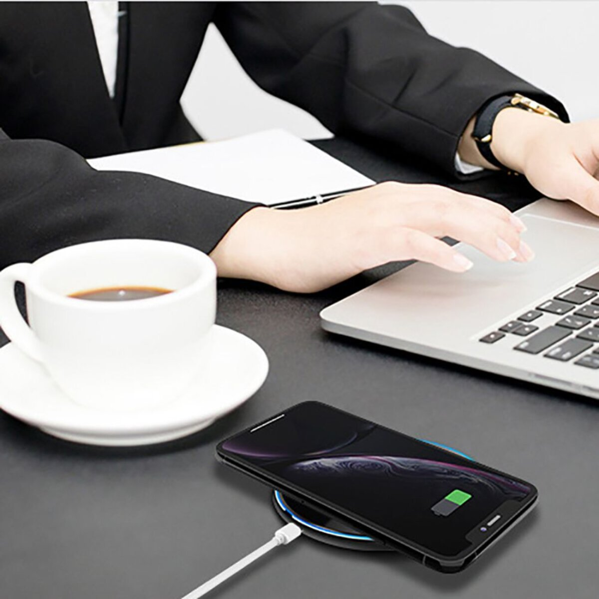 Eine Person benutzt einen Laptop. Ein Smartphone wird drahtlos auf einem Pad aufgeladen. Eine Tasse Kaffee und Untertasse stehen auf dem Schreibtisch.
