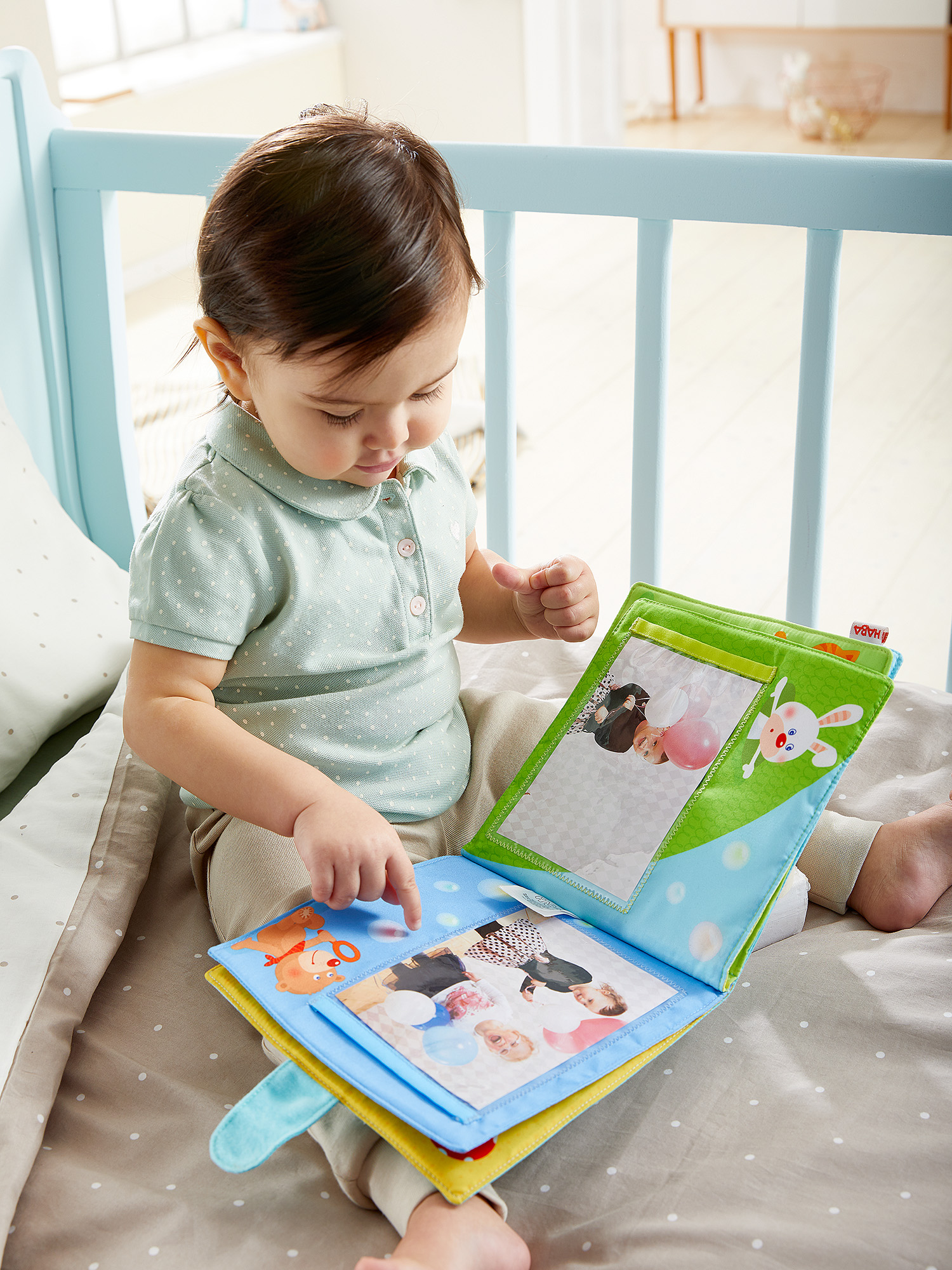 Ein Baby sitzt in einem blauen Kinderbett und betrachtet ein buntes Stoffbuch mit Fotos. Der Hintergrund zeigt einen Raum.