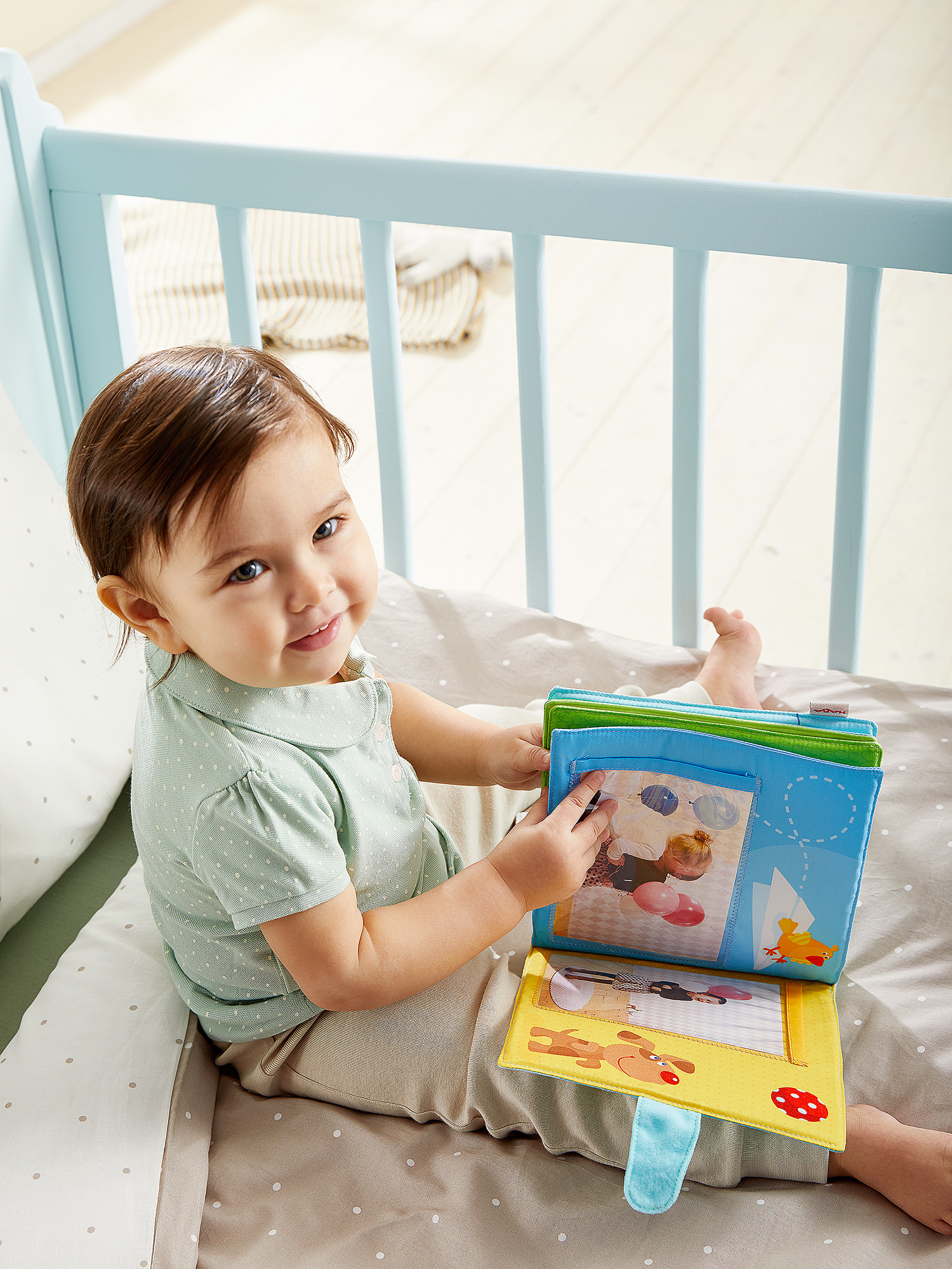 Ein lächelndes Baby hält ein farbenfrohes Buch in einem Kinderbett. Das Buch zeigt Bilder von Spielzeug und Tieren. Das Baby trägt ein grünes Hemd.