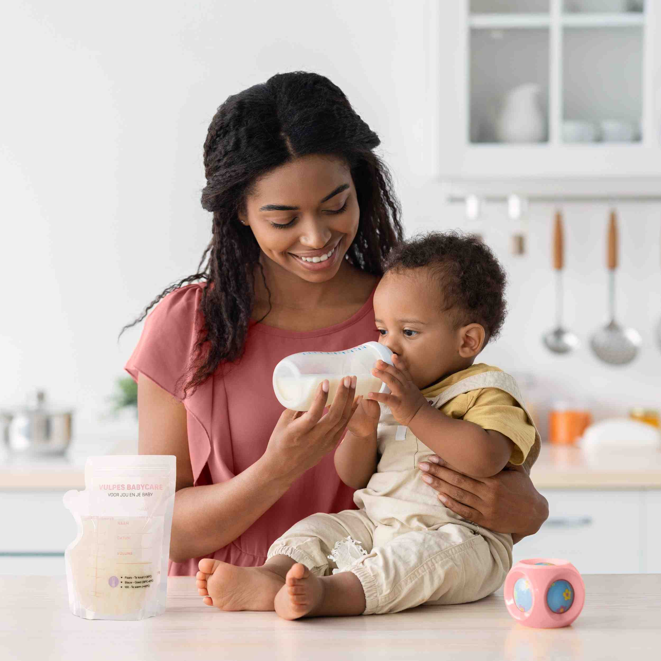 Een vrouw voedt een baby met een fles. Een melkzak en een speeltje liggen op tafel.