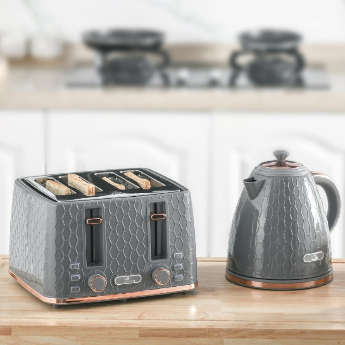 Ein grauer Toaster mit Toast und ein grauer Wasserkocher mit kupferfarbenen Akzenten stehen auf einer Holzoberfläche. Im Hintergrund ist eine Küche zu sehen.