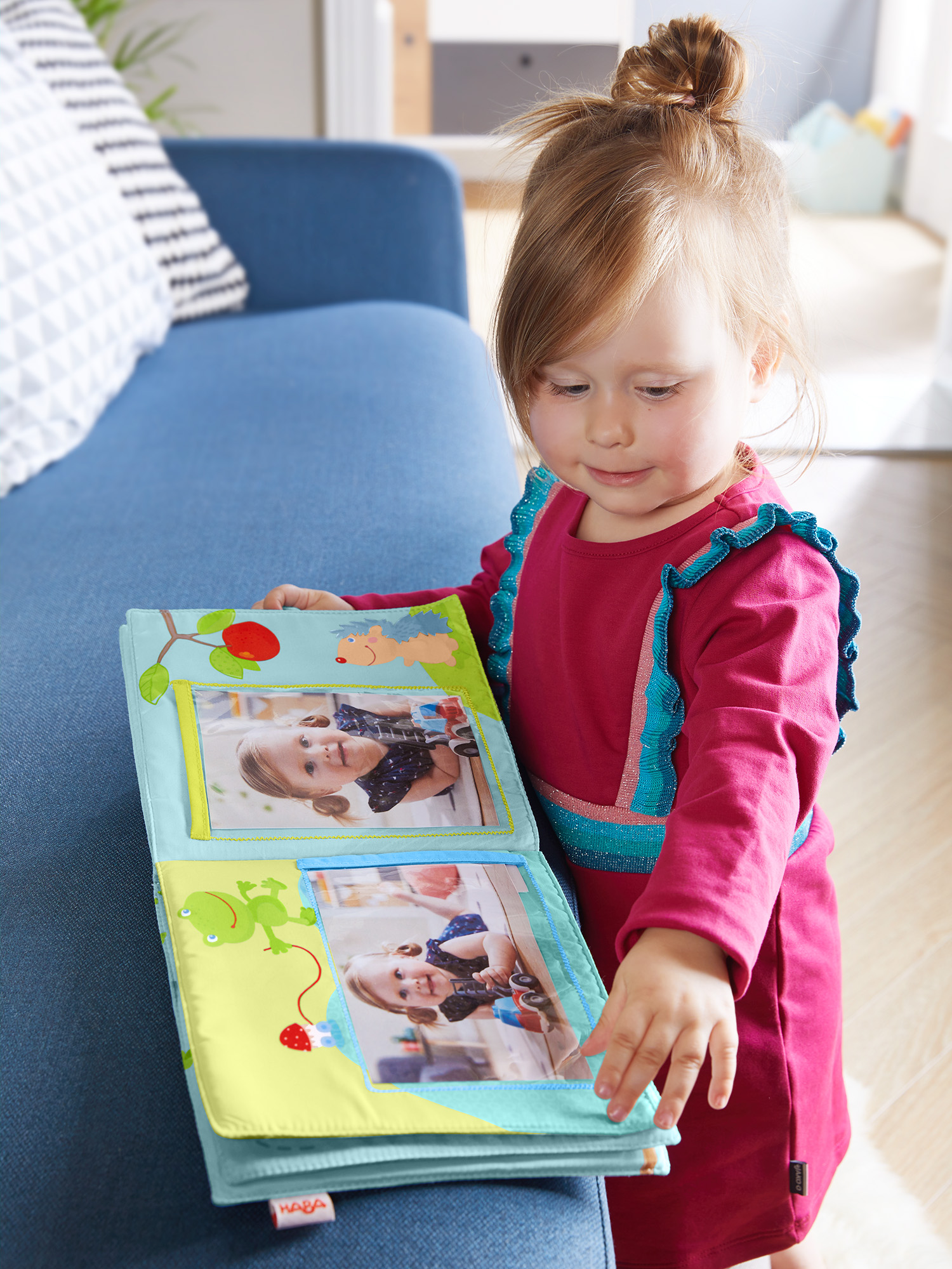 Ein kleines Mädchen in einem roten Kleid betrachtet ein Fotoalbum, blaue Couch im Hintergrund.