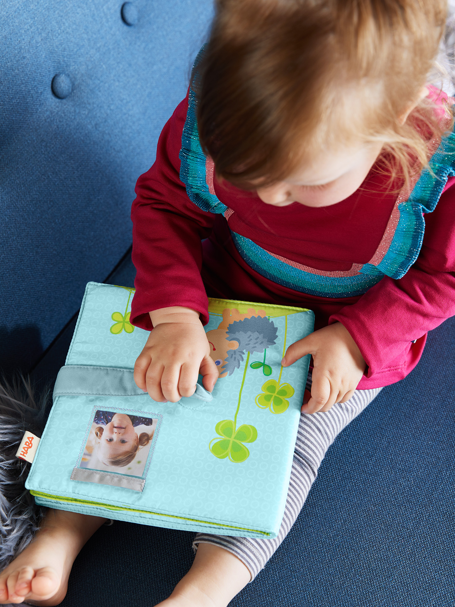 Ein Kind in einem roten Hemd hält ein blaues Buch auf einem blauen Stuhl. Das Buch zeigt ein Bild eines Kindes.