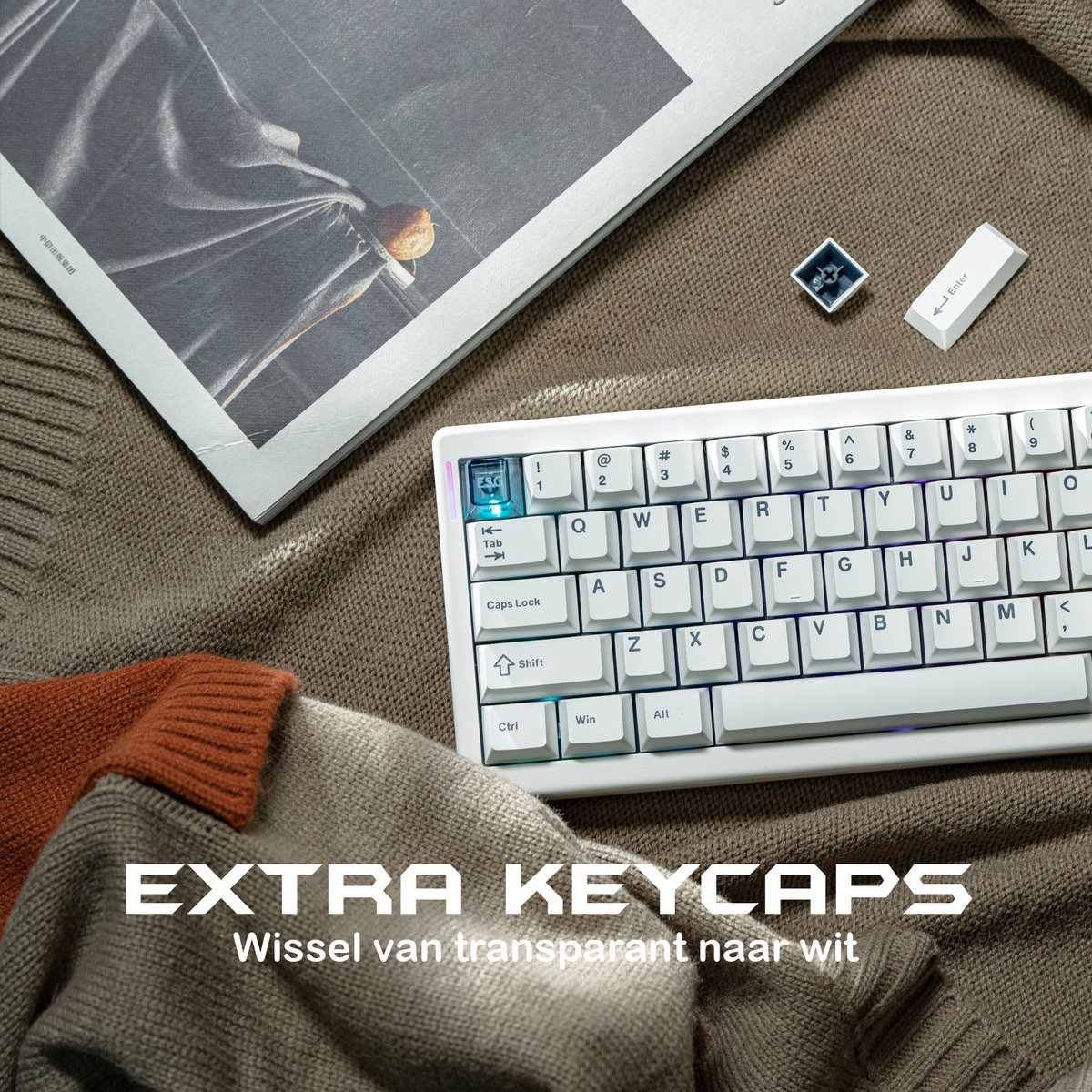 A white keyboard with extra keycaps is on a brown blanket. A magazine and two sweaters are also in the frame.