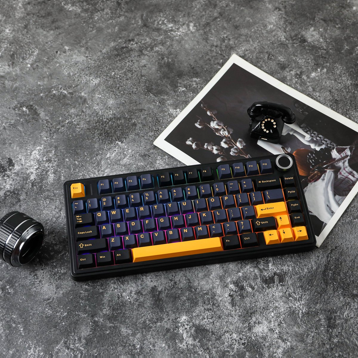Eine schwarze mechanische Tastatur mit gelben und orangen Tasten liegt auf einer grauen Oberfläche. Ein Vintage-Telefon und ein Foto sind im Hintergrund.