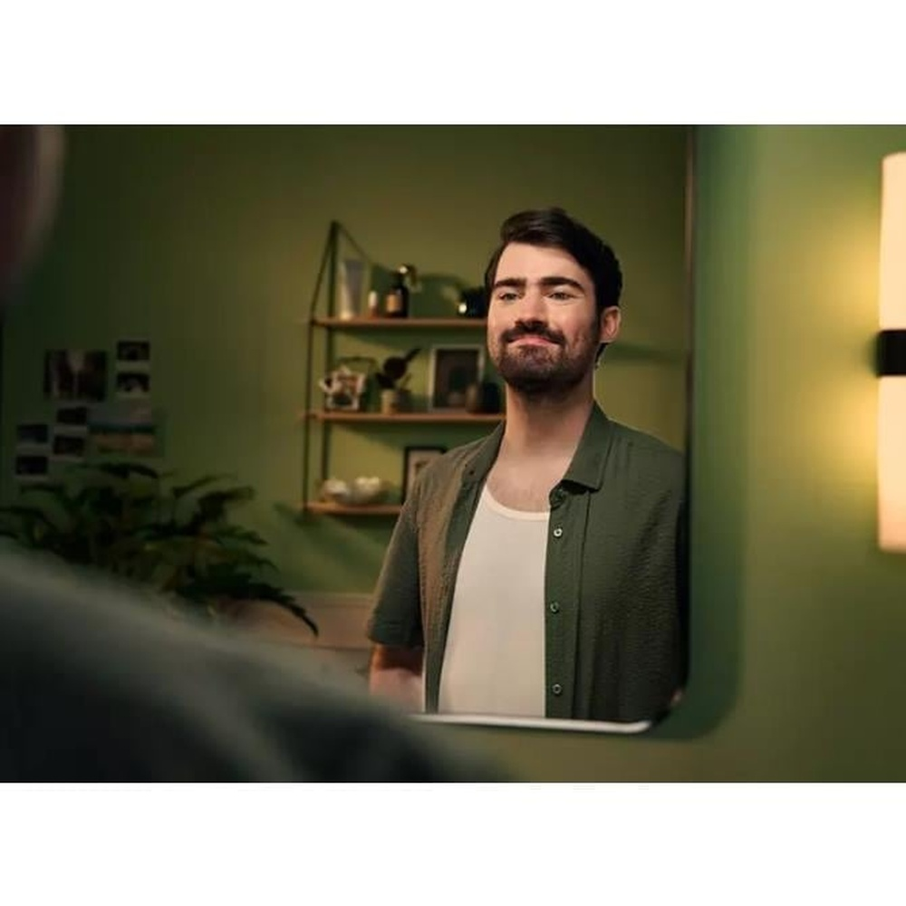 Mężczyzna patrzy w lustro. Ma na sobie zieloną koszulę na białej koszulce. W tle półka z przedmiotami.