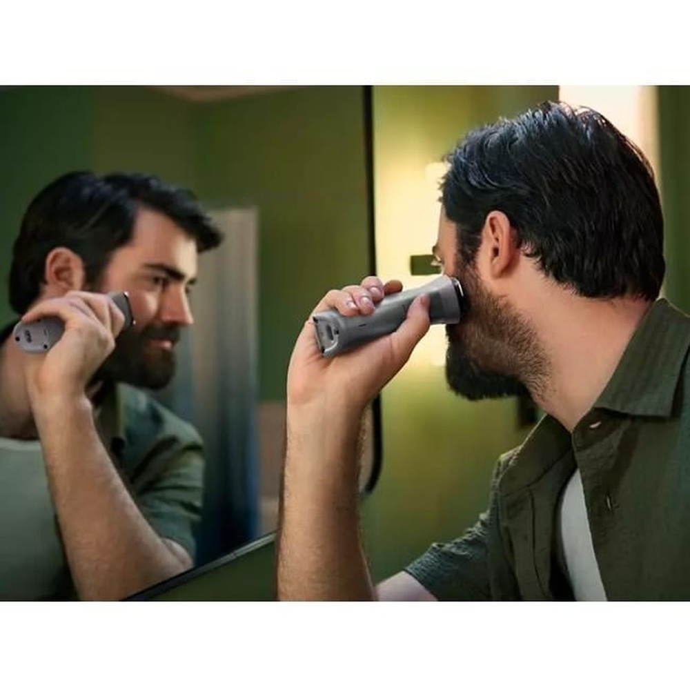 Mężczyzna goli brodę przed lustrem trymerem. Lustro odbija mężczyznę i trymer.