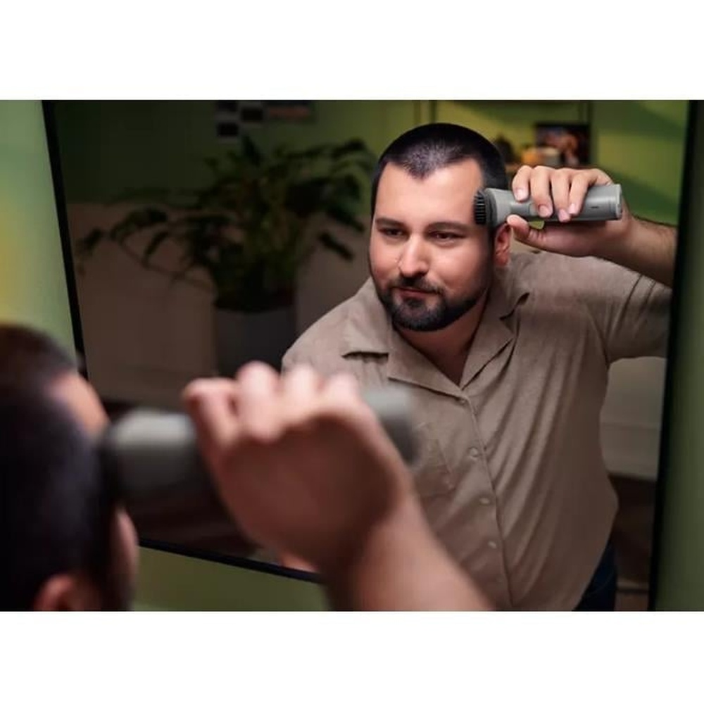 Mężczyzna goli głowę elektryczną maszynką do strzyżenia przed lustrem. Ma na sobie beżową koszulę.