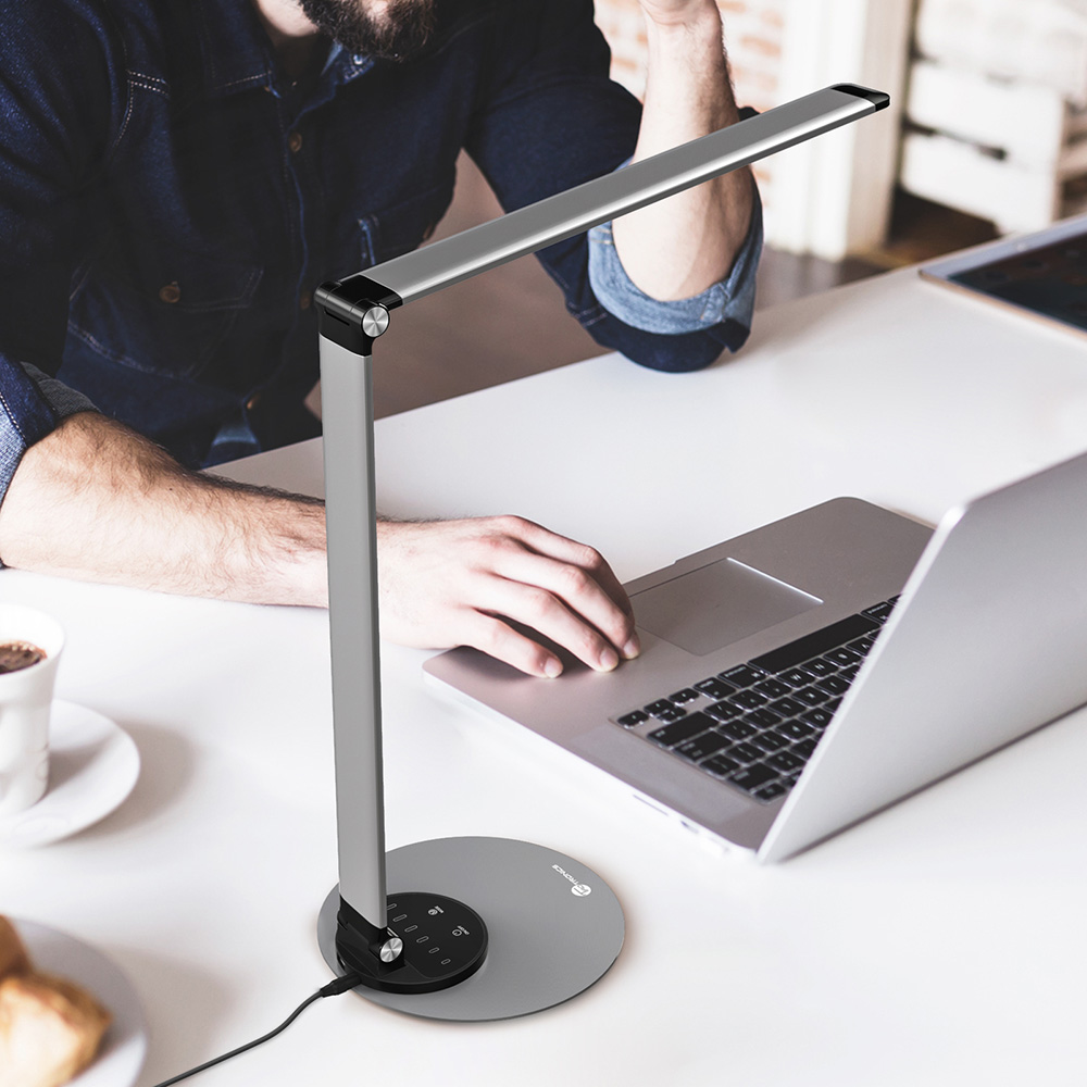 Eine Person benutzt einen Laptop, links steht eine moderne graue Schreibtischlampe. Kaffee und Brot stehen auch auf dem weißen Schreibtisch.